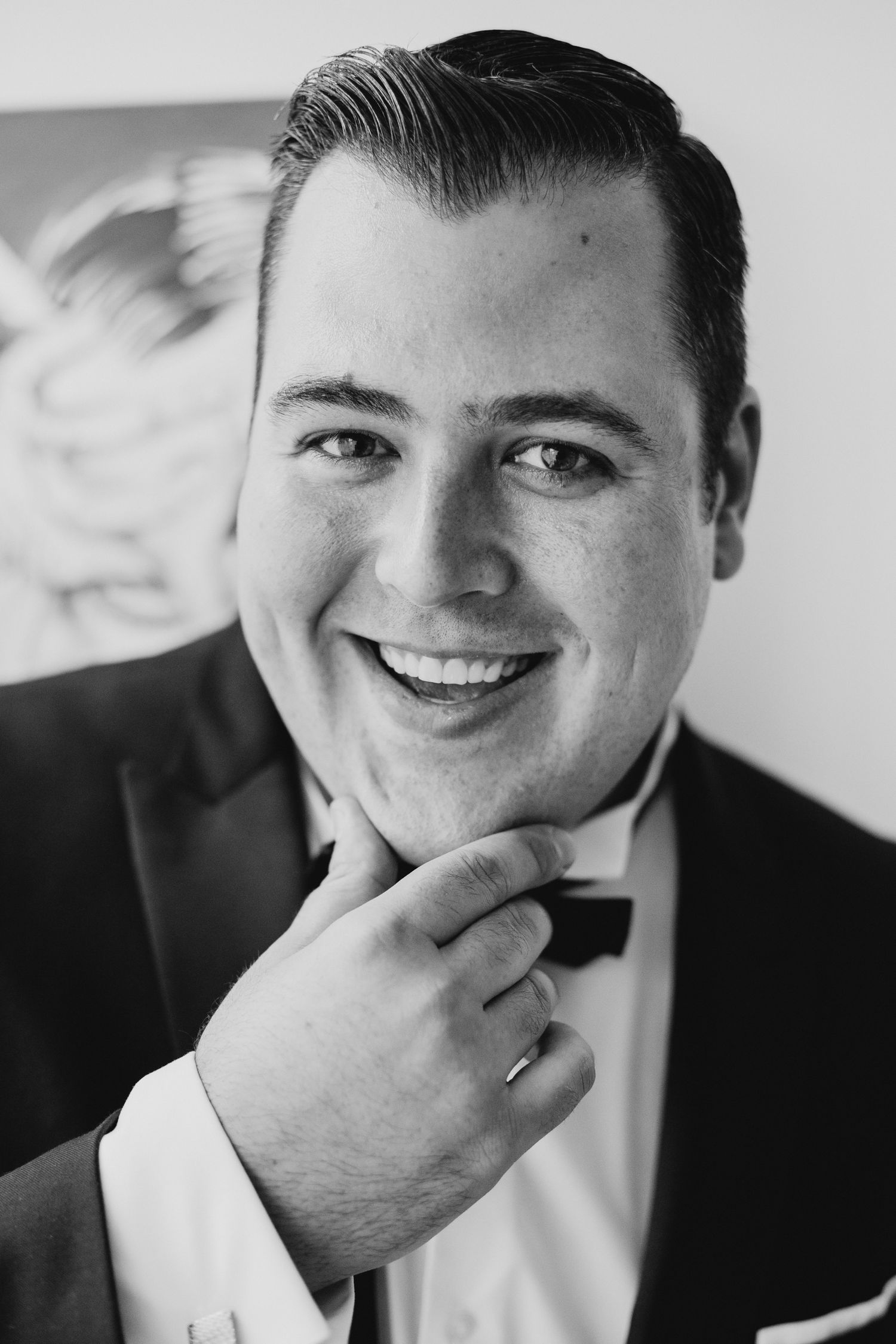 Black and white portrait of person in formal attire with bow tie smiling while touching their chin.