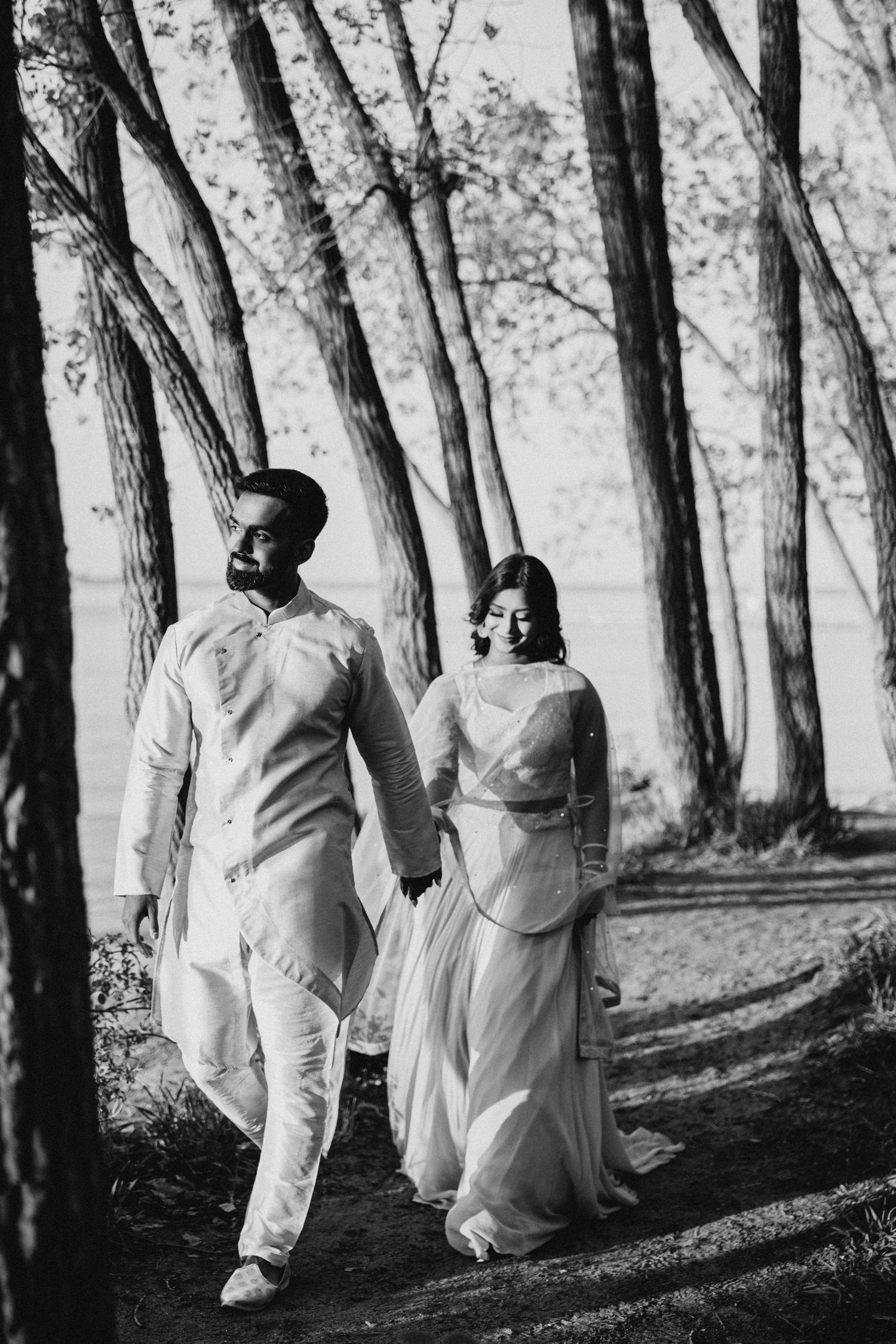 A black and white photo of a couple walking hand in hand through a tree-lined path.