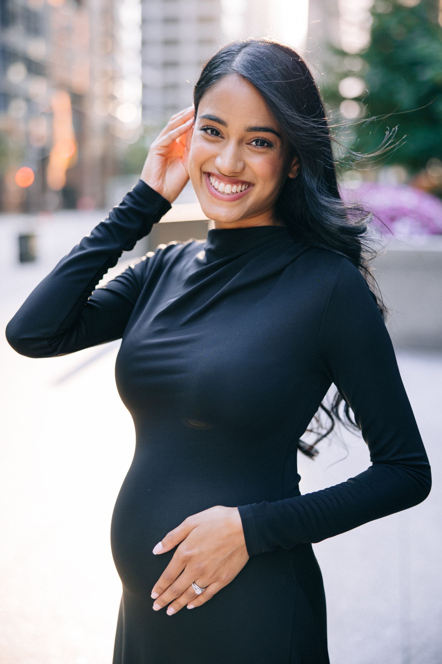 A smiling pregnant person in a fitted black dress touches their hair against a blurred urban backdrop.