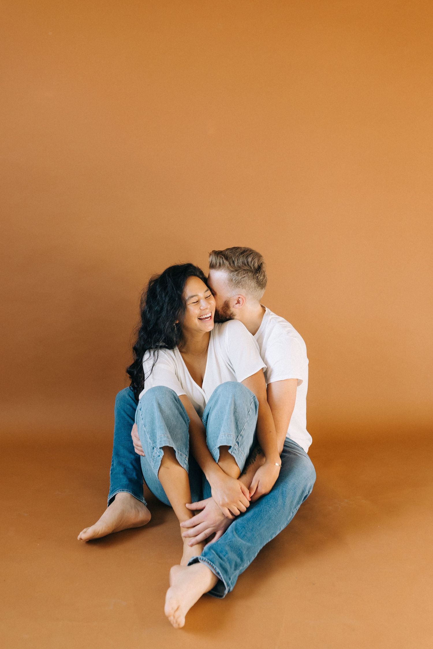A couple in casual white shirts and jeans sits intimately on a brown backdrop sharing a joyful moment together.