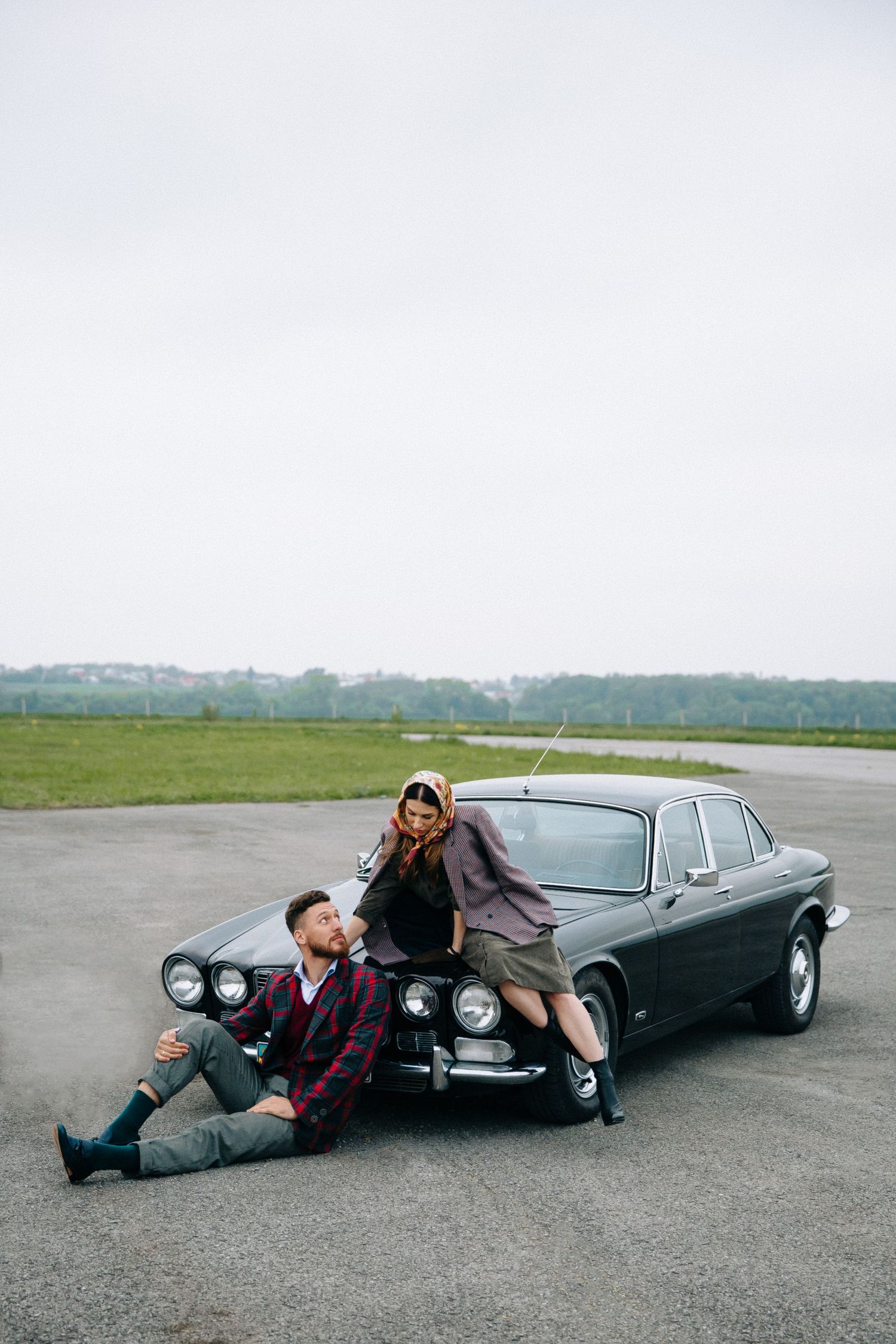 Two people in casual attire pose with a vintage black car on an empty airfield on an overcast day.
