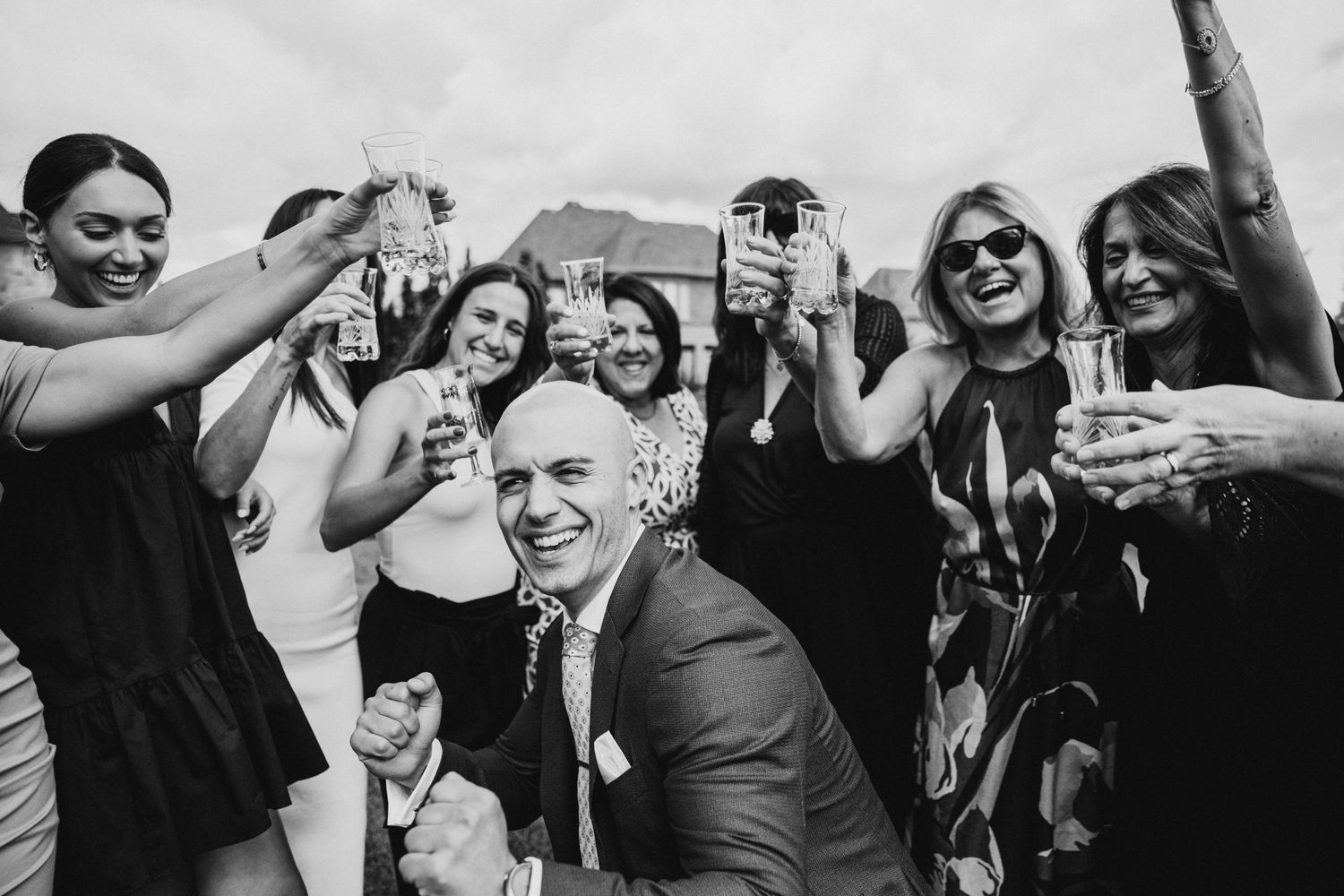 A black and white photograph showing a joyous wedding celebration with people raising glasses in a festive toast.