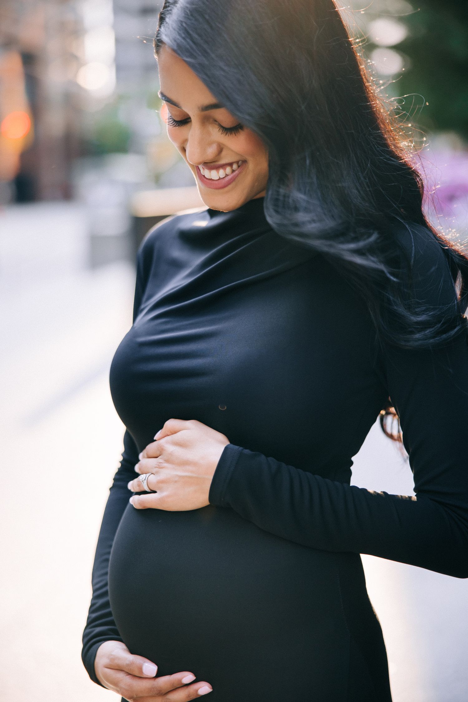 Glowing pregnant woman in black dress holding belly while smiling at urban setting.