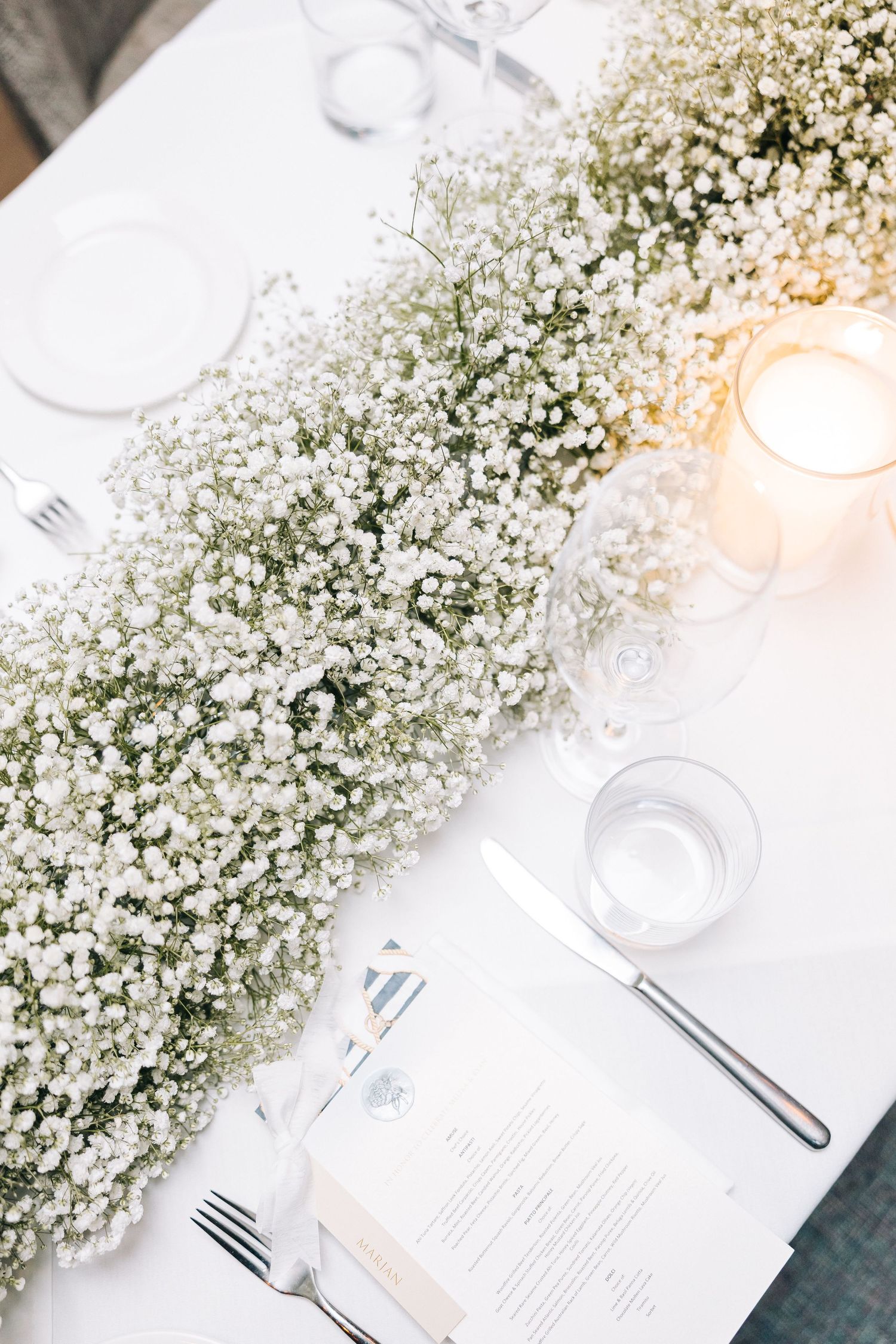 Elegant table setting with baby's breath floral runner, white linens, glowing candles and place settings with menu cards.
