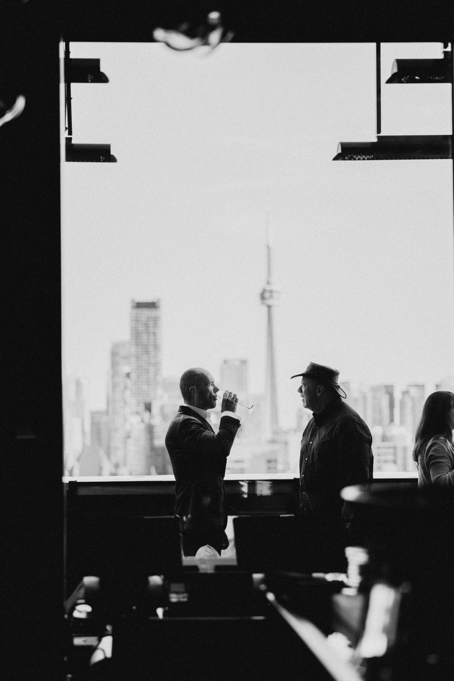 Two patrons converse at a bar with the silhouette of Toronto's CN Tower and skyline visible through a window.