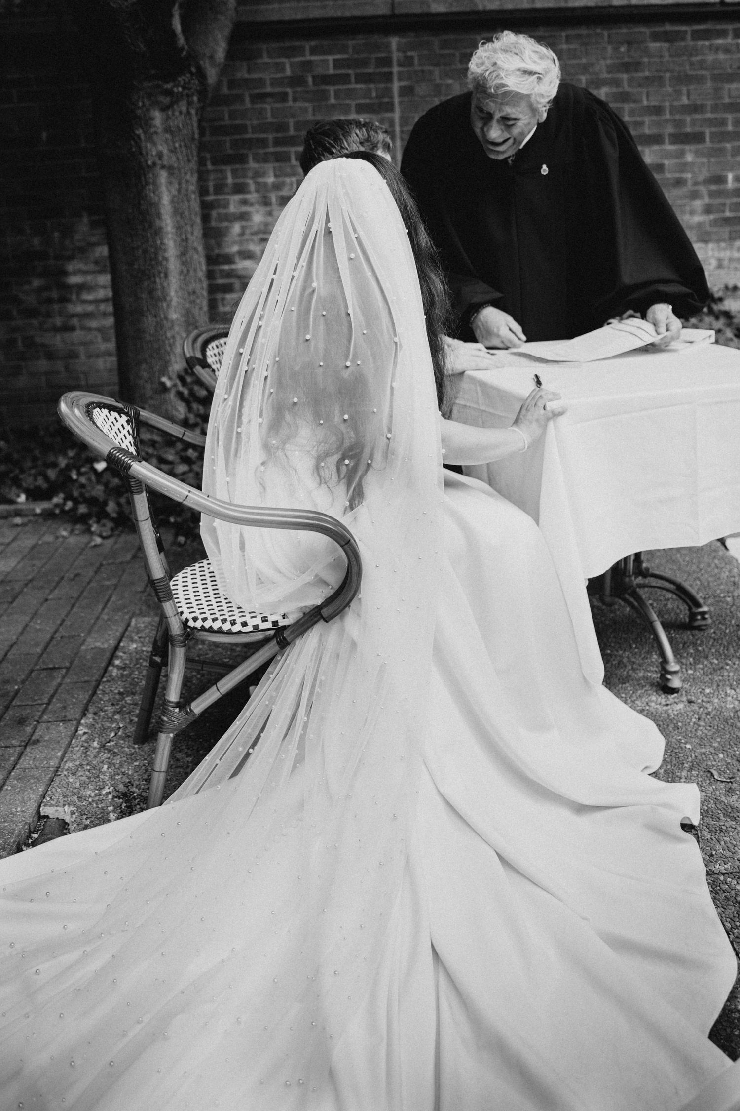 A bride in a flowing veil signs documents with an officiant at an outdoor ceremony.