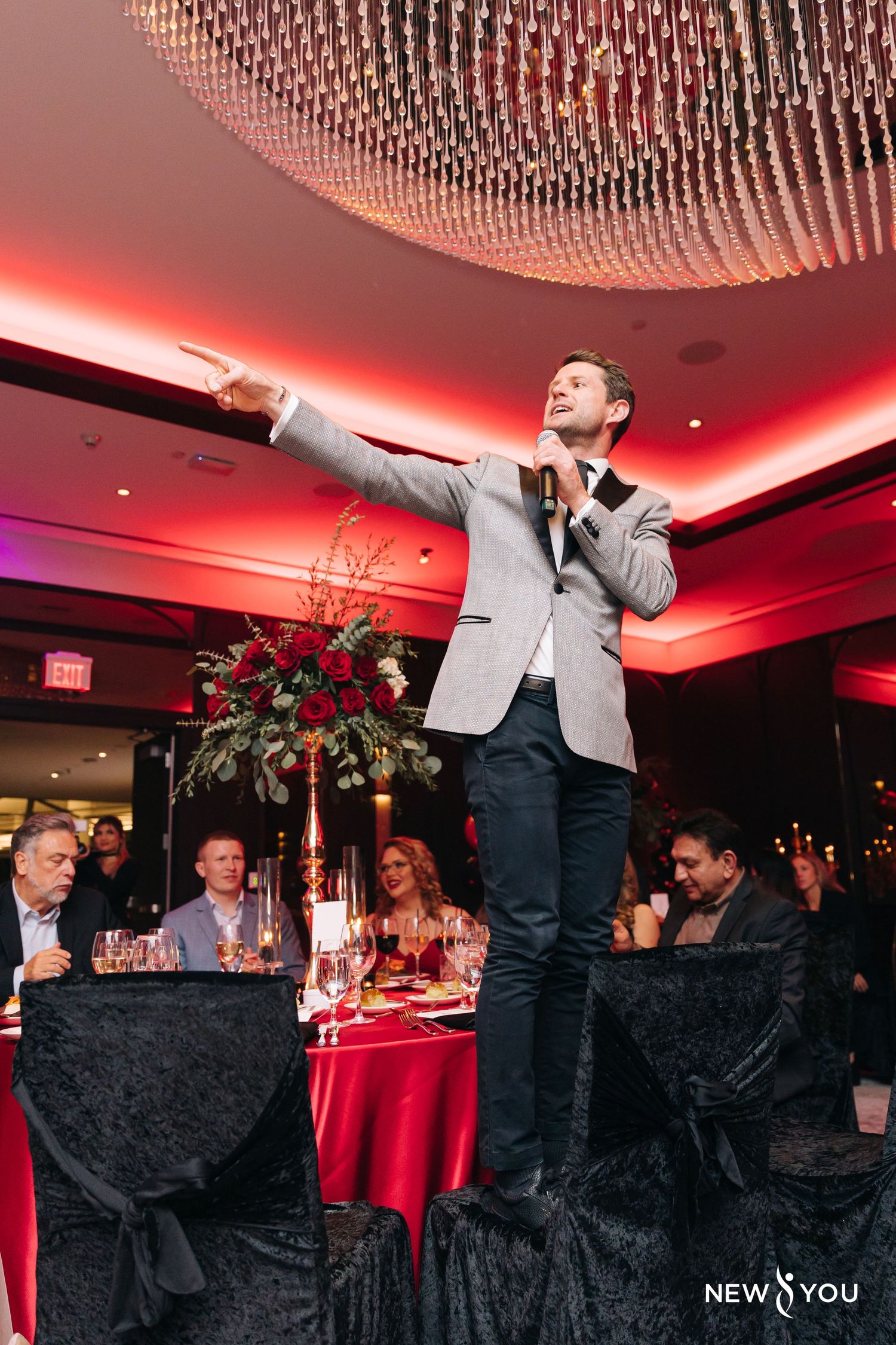 Speaker in grey blazer addressing audience at formal dinner event with red ambient lighting.