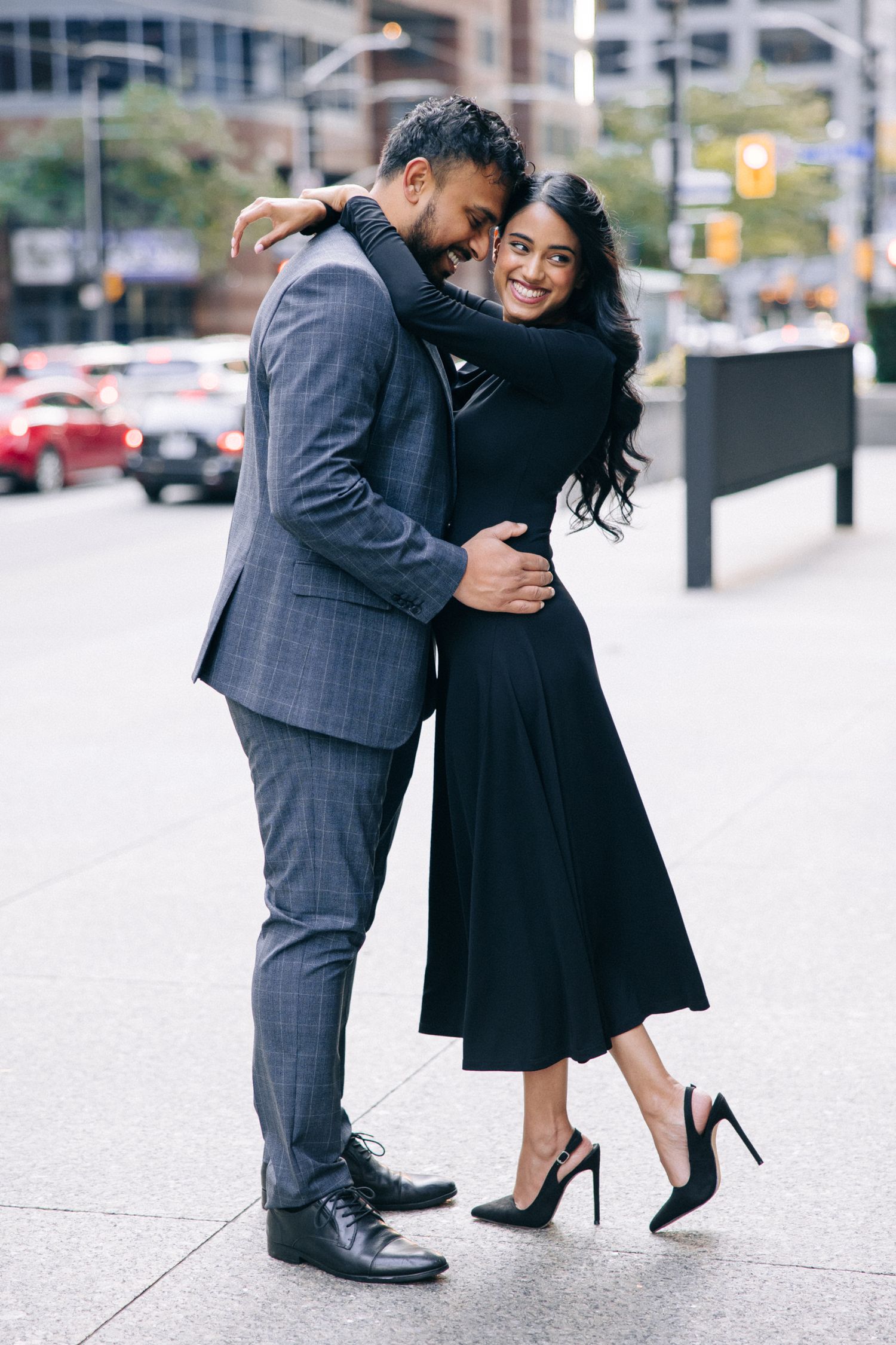 A couple embraces on a city street, with the man in a grey suit and the woman wearing a flowing black dress and heels.