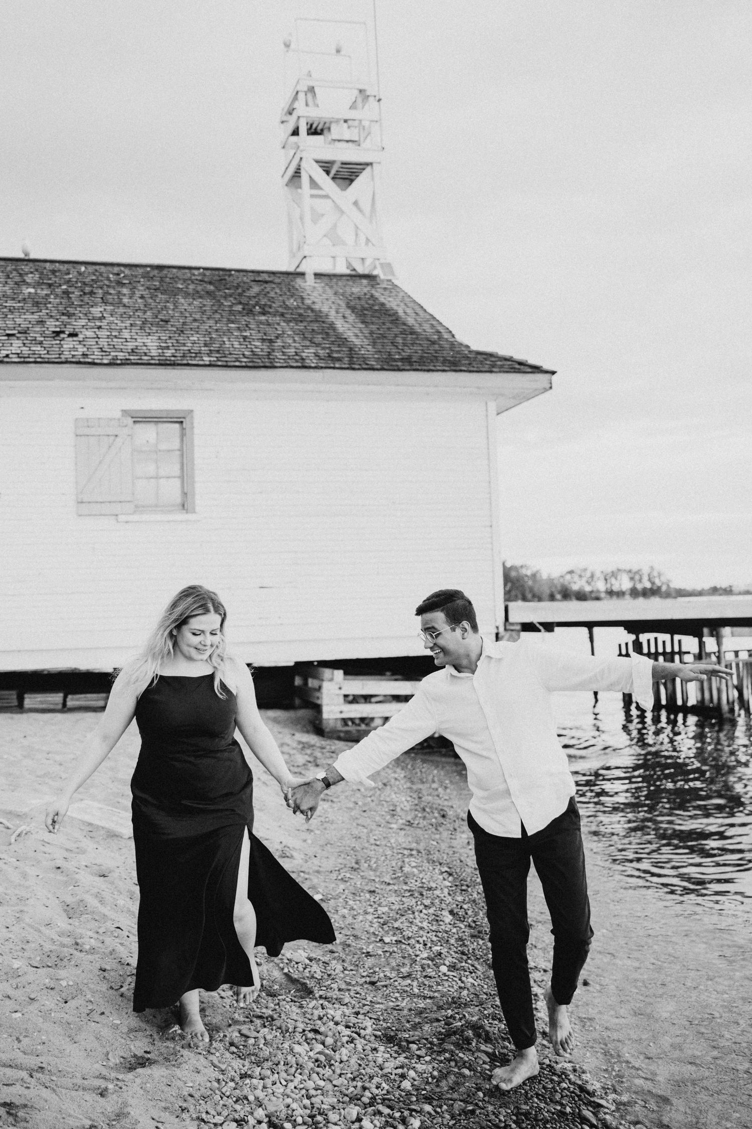 A couple holds hands and dances by a lighthouse on the shore.