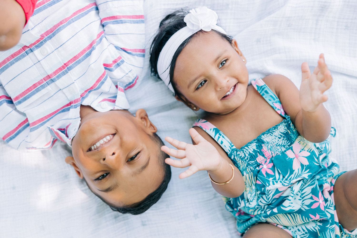 Two children in summer outfits lying on white blanket smiling together.