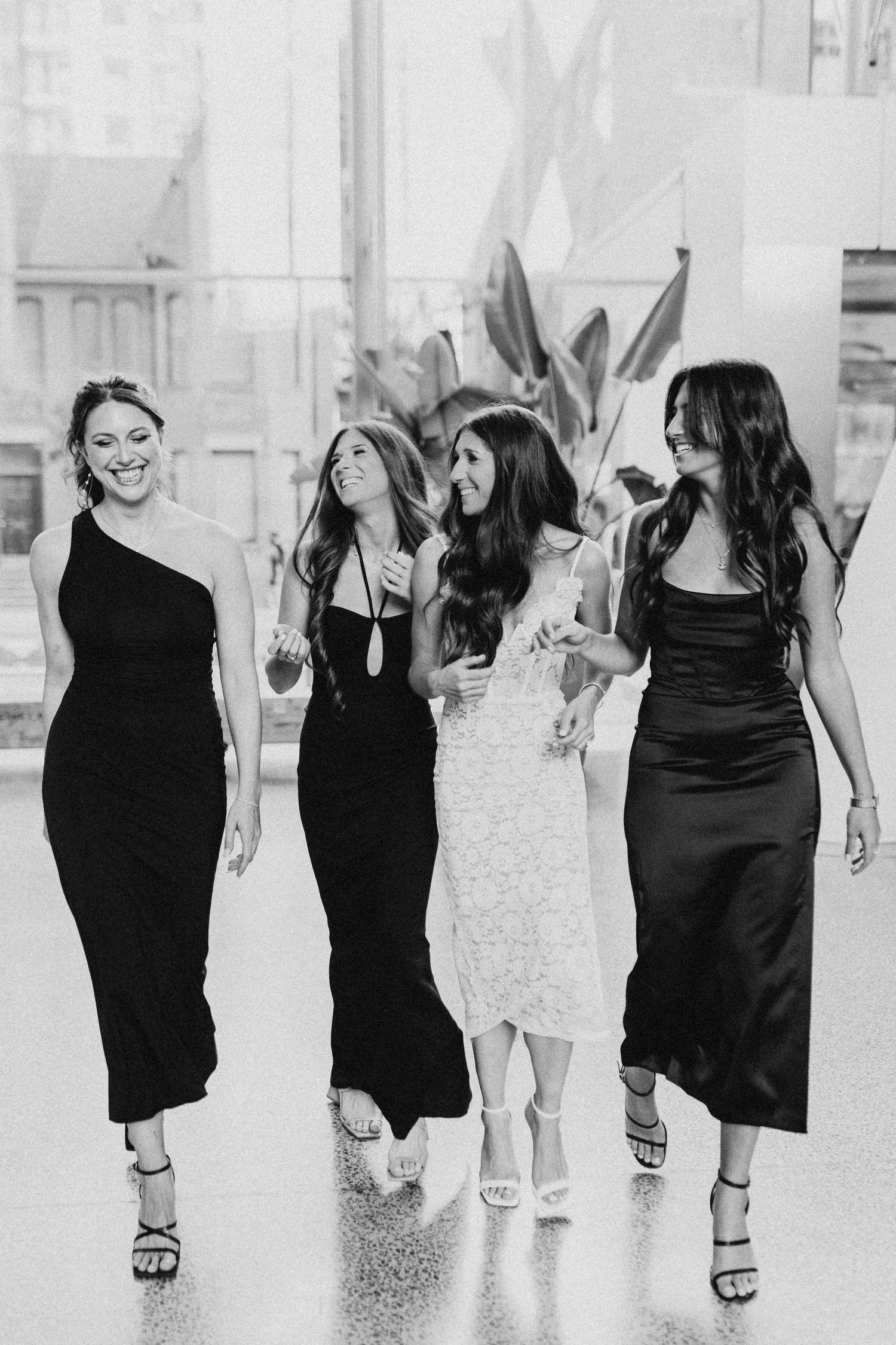 Four women in black dresses share joyful moment in bright indoor setting.