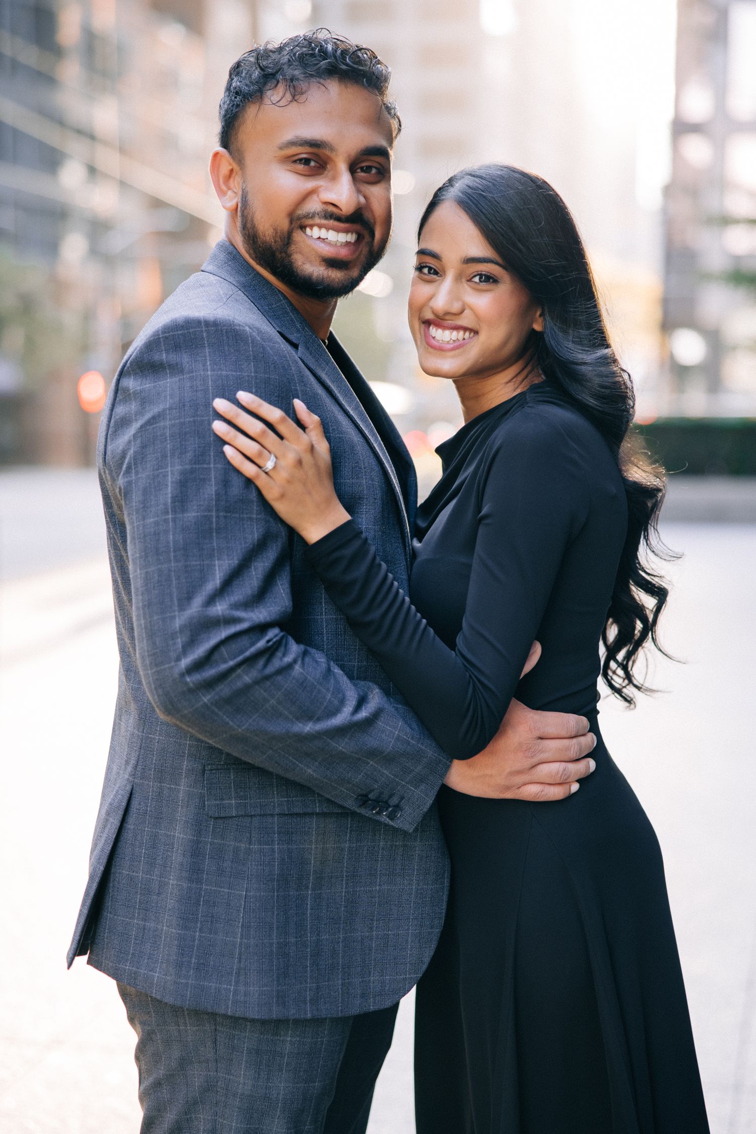 A couple embraces while dressed elegantly in black and grey formal attire against an urban backdrop.