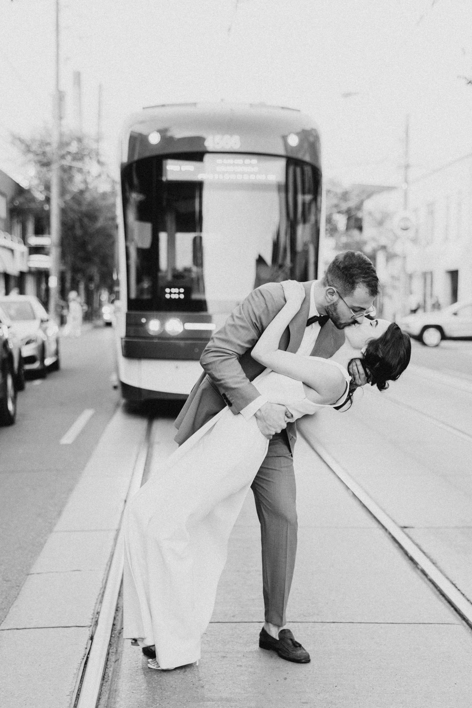 A romantic couple share an intimate dip kiss in front of a streetcar on an urban street.