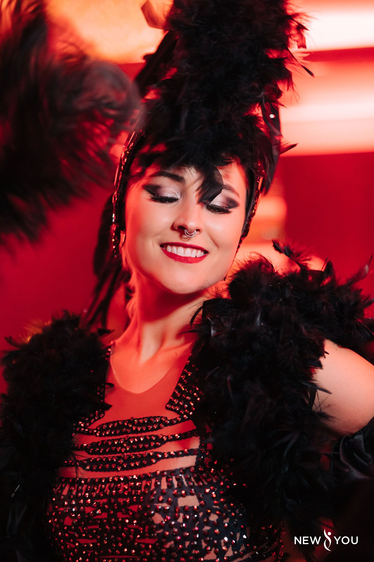 Burlesque performer in black feathered headpiece and sparkly costume smiling under red lights.