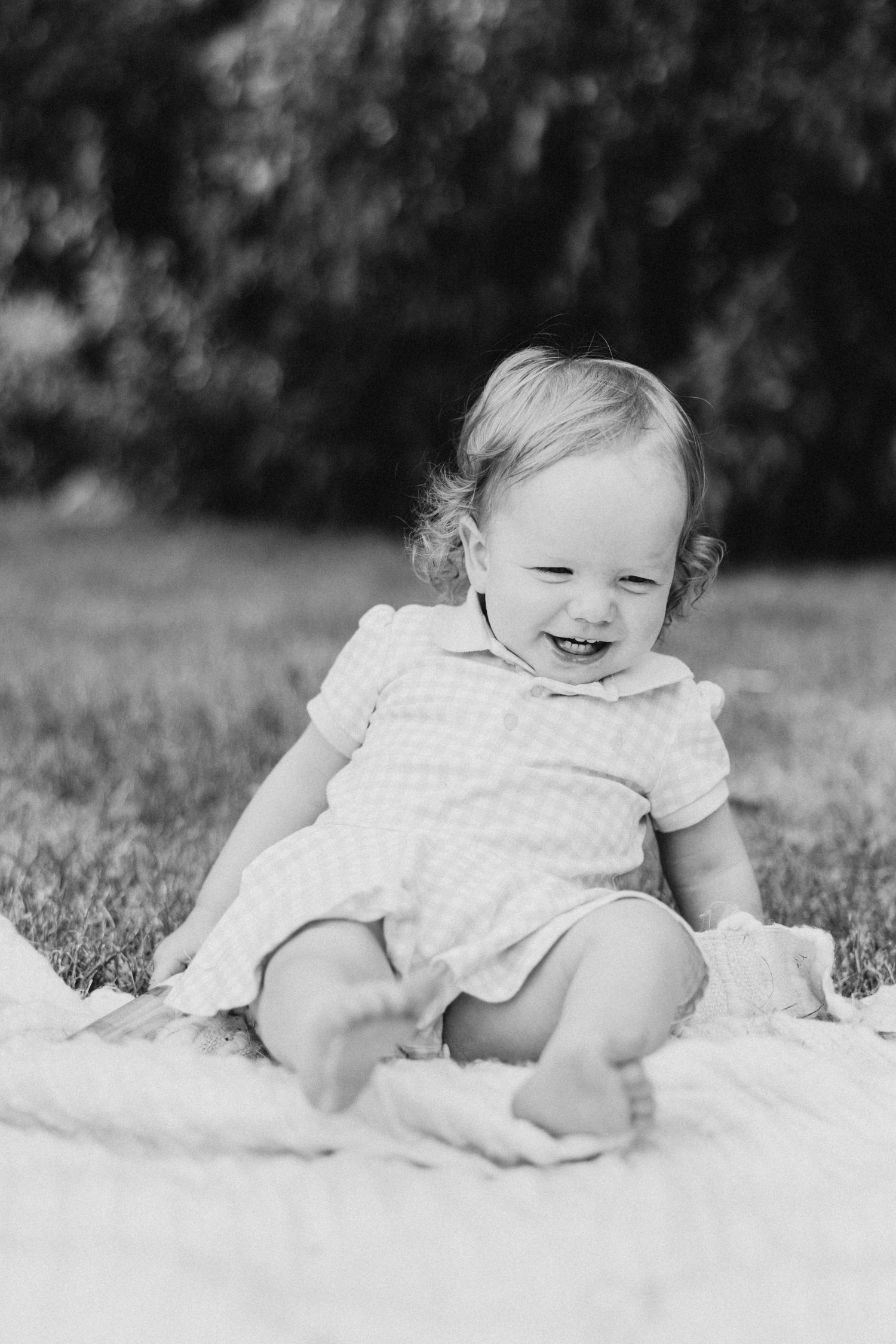Baby in gingham dress sitting on white blanket outdoors in black and white portrait.