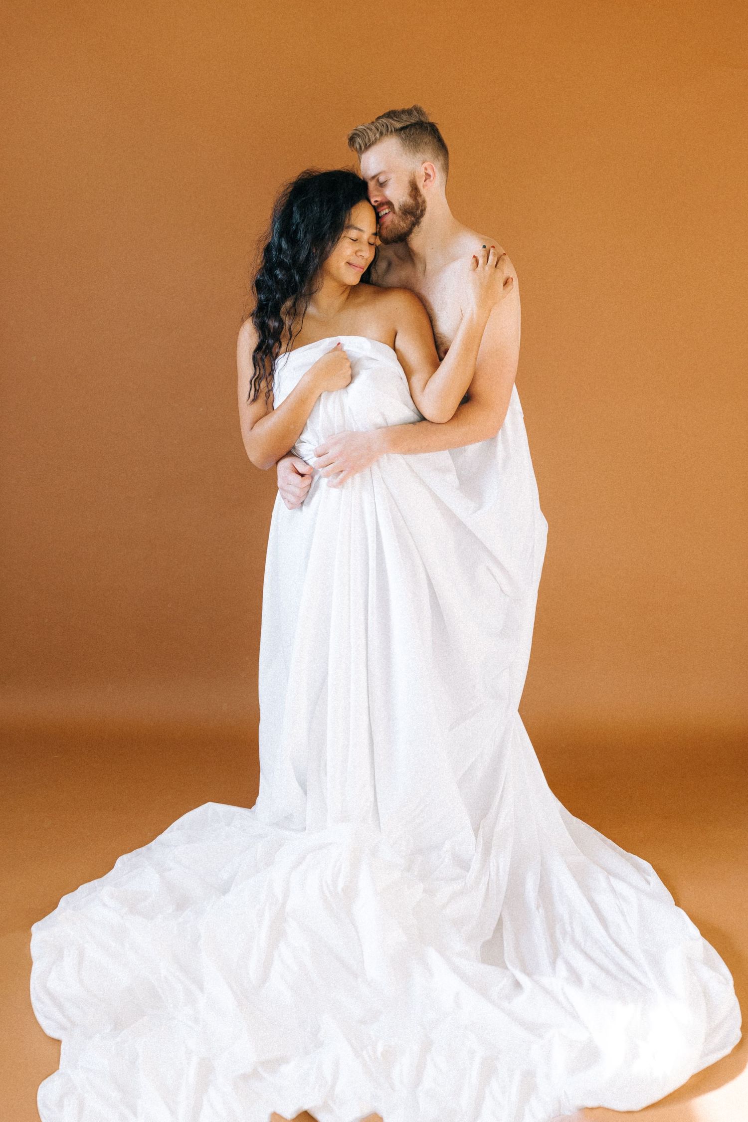 Romantic couple wrapped in flowing white fabric against terracotta background.