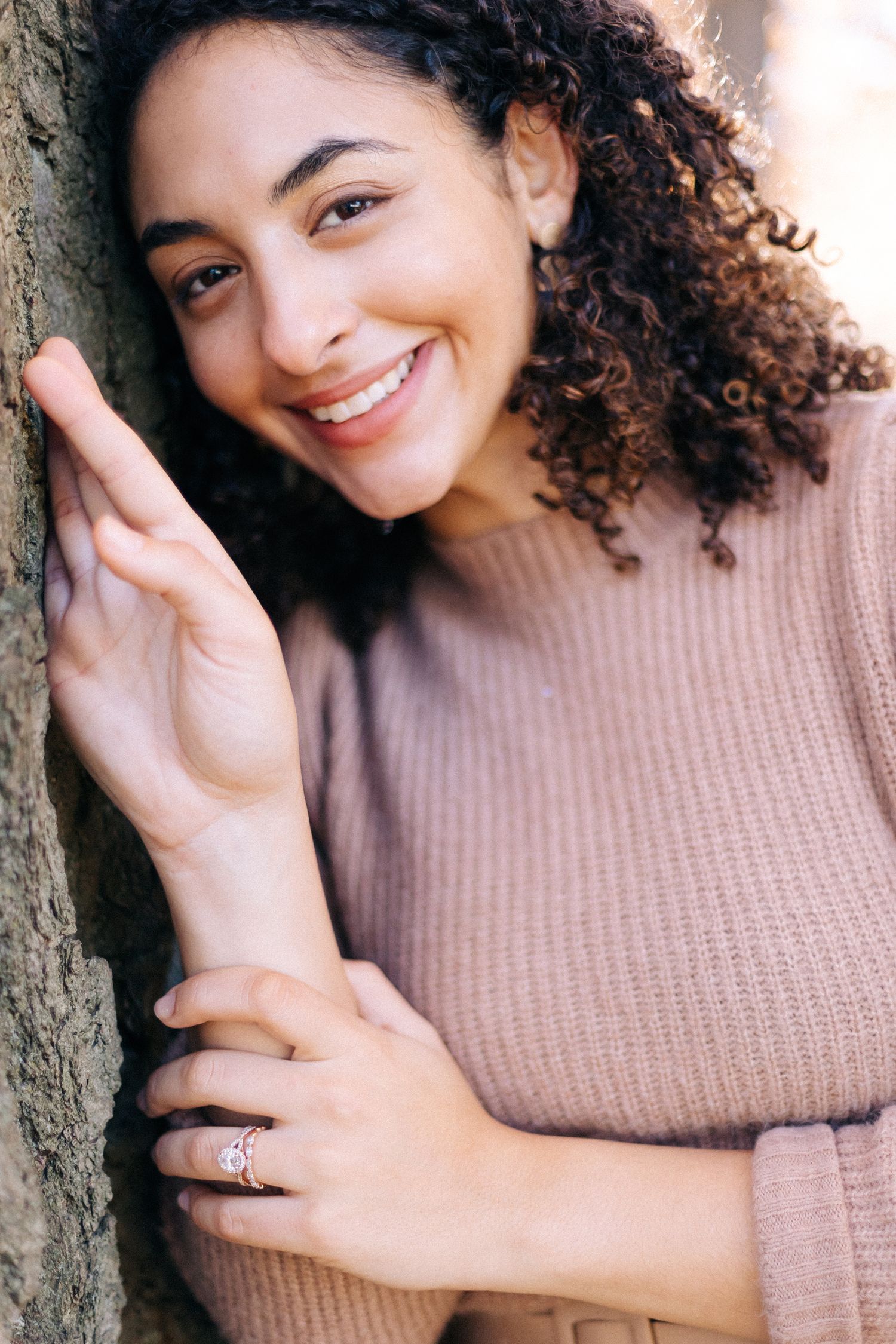 A person with curly hair smiles warmly while leaning against a tree.