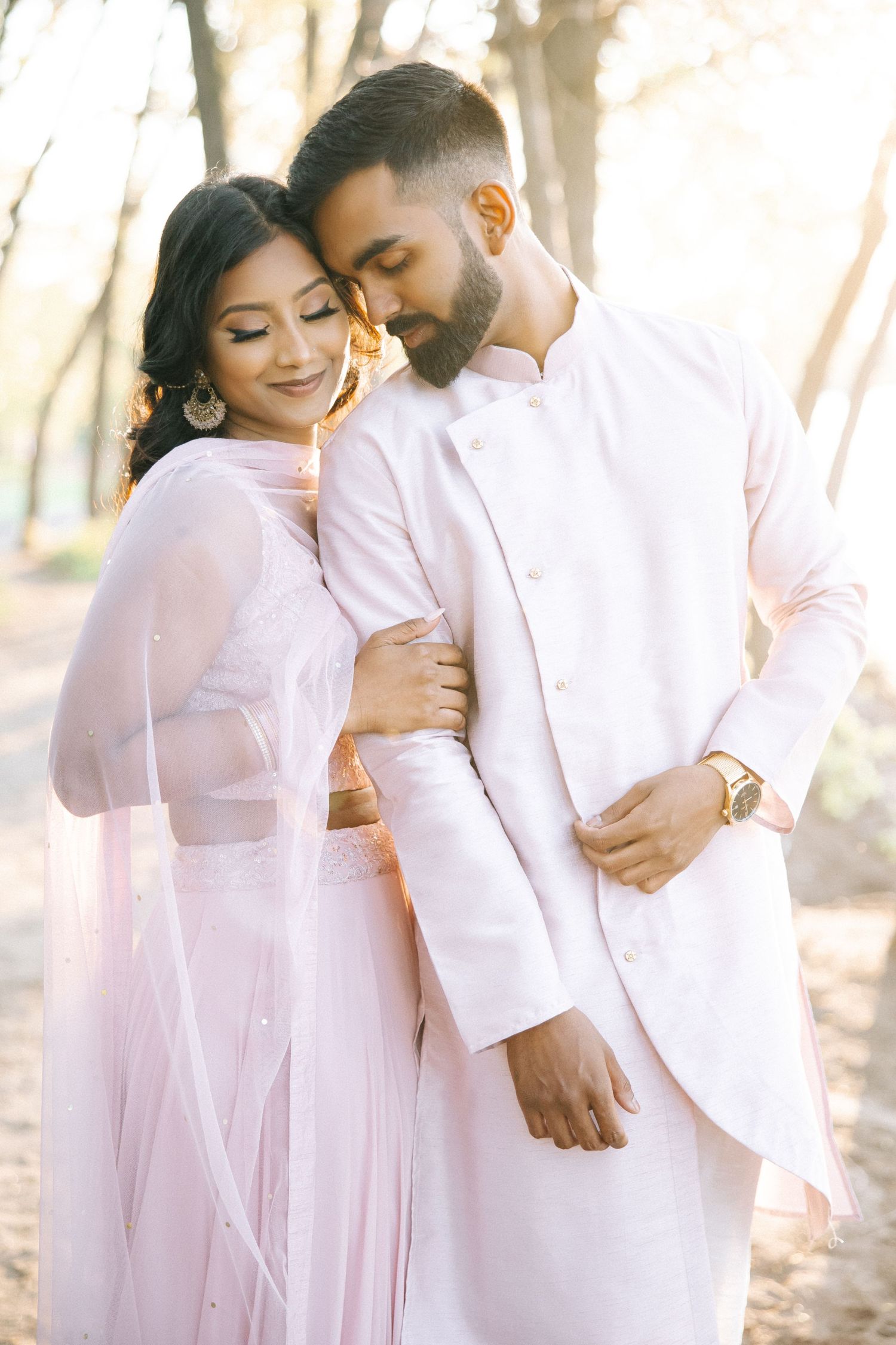 A couple in soft pink and white traditional outfits poses together in a sunlit forest setting.
