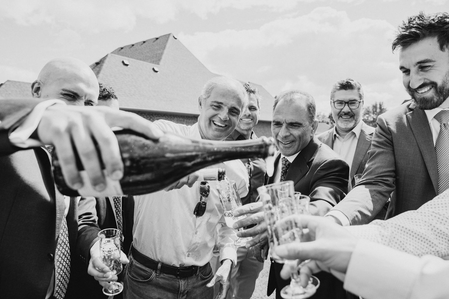 Black and white photo showing people celebrating with champagne being poured into glasses outdoors.