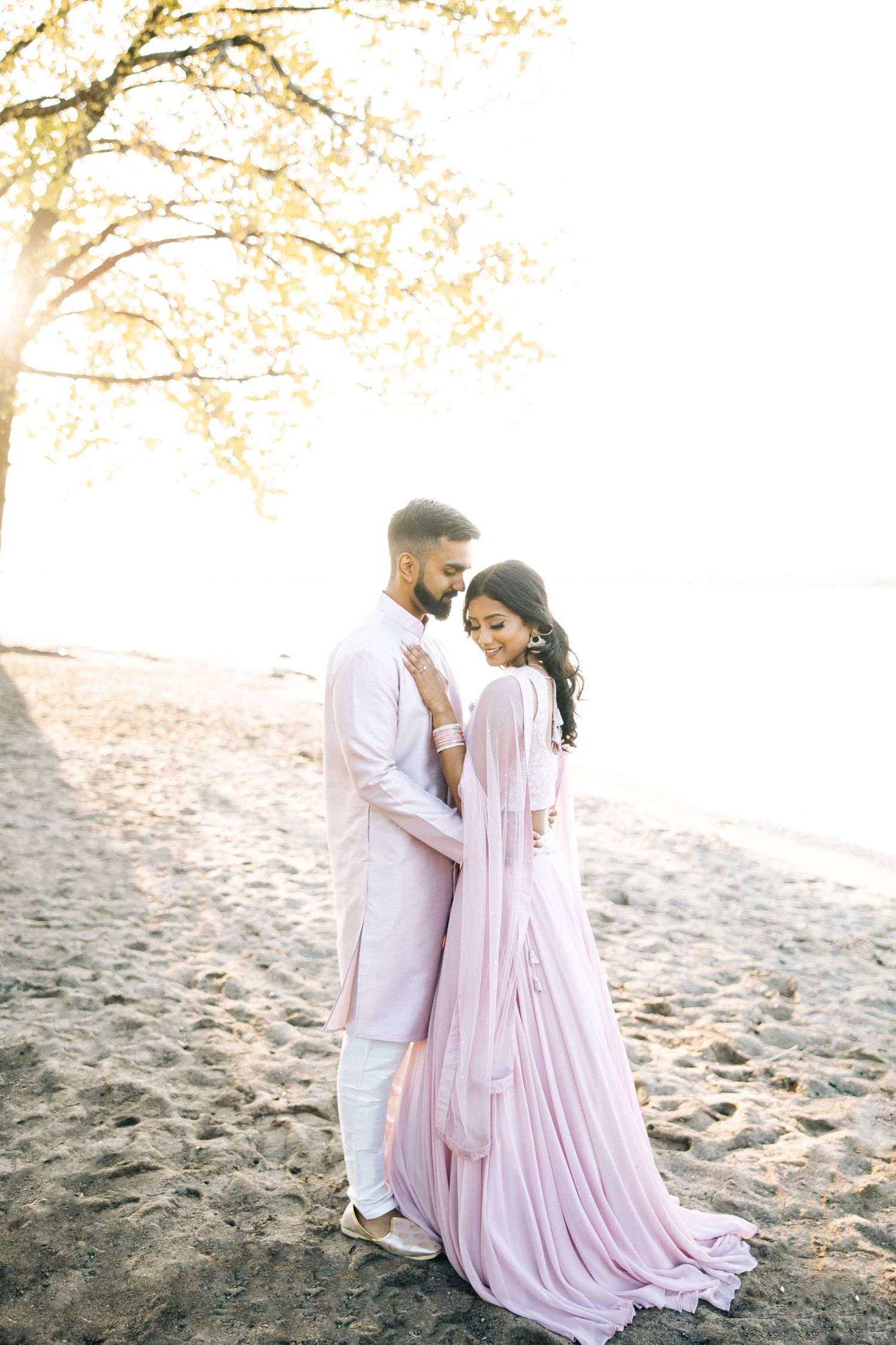 Romantic couple in white and blush outfits embrace on sandy beach during sunset photo session.