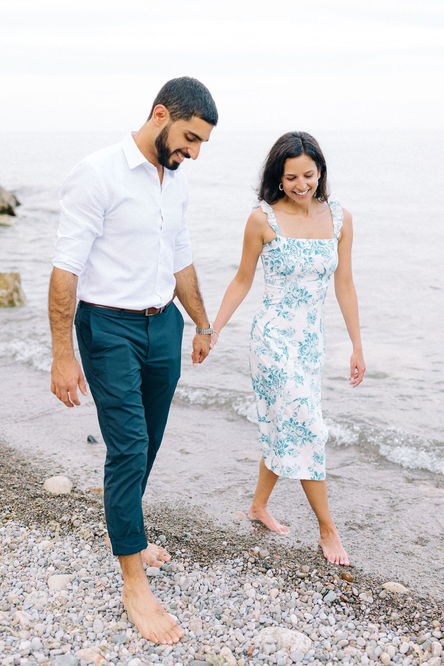 A couple walks barefoot along a rocky beach shore.