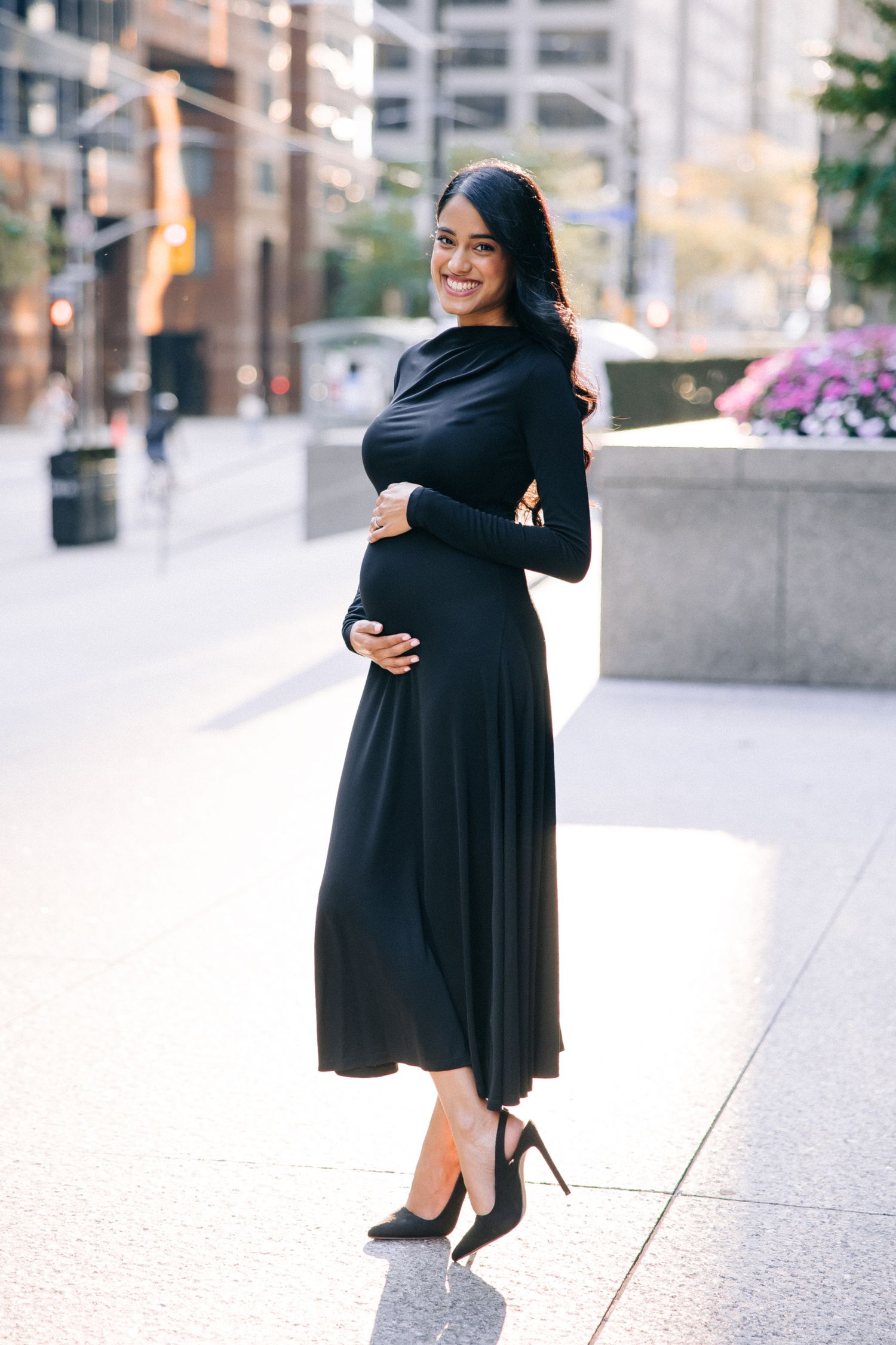 A pregnant woman in a flowing black dress and heels poses elegantly on a city sidewalk with buildings in the background.