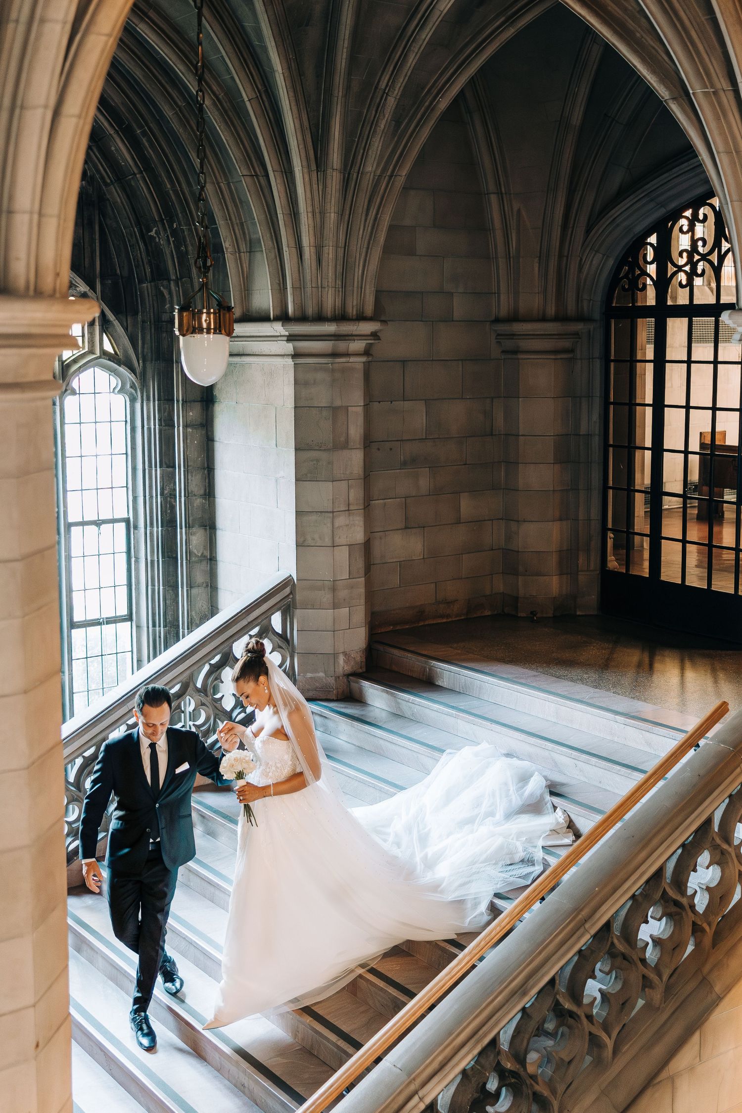 A couple ascends an elegant stone staircase in a Gothic-style building with arched windows and vaulted ceilings.