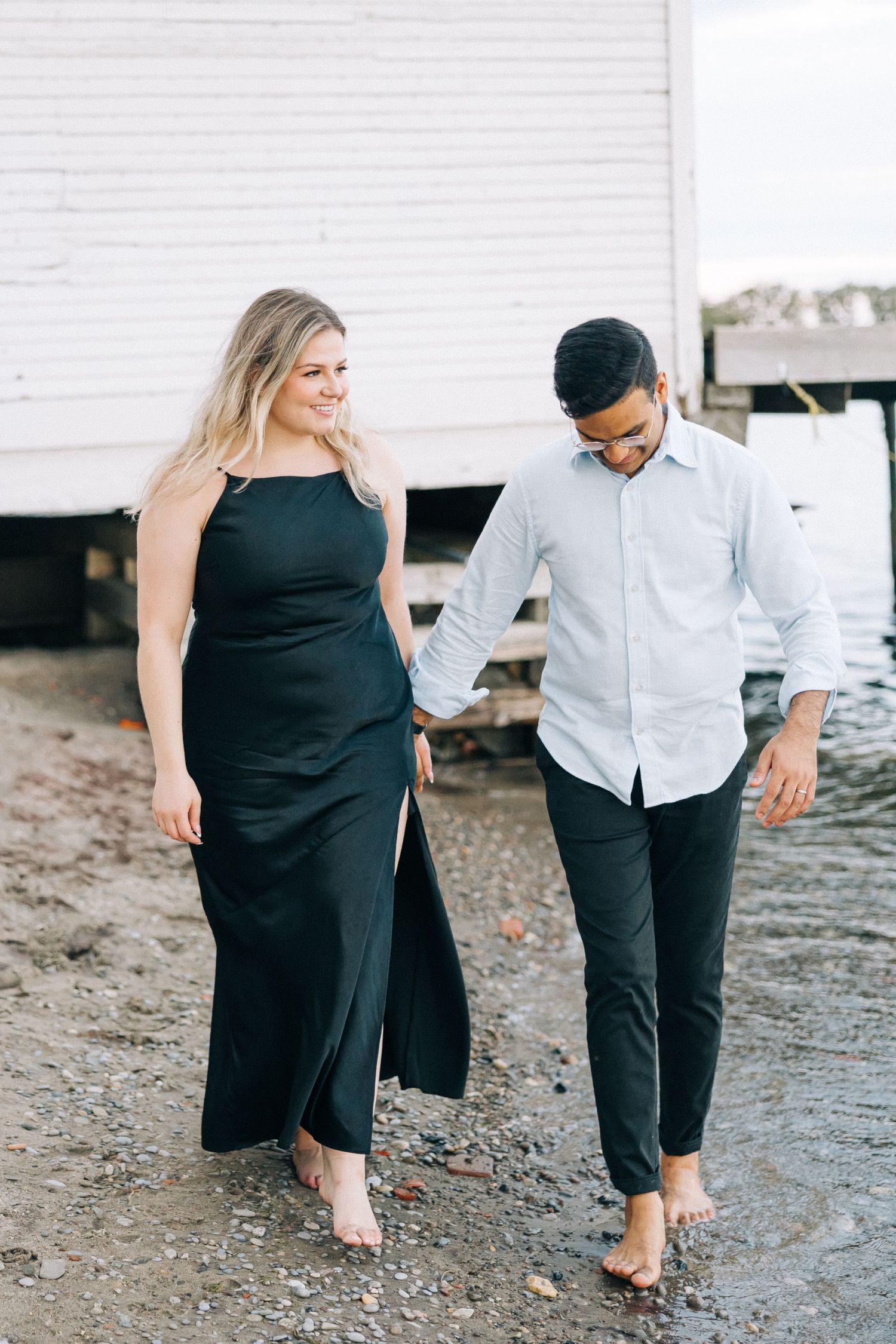 A couple walks barefoot along a beach holding hands in formal attire.
