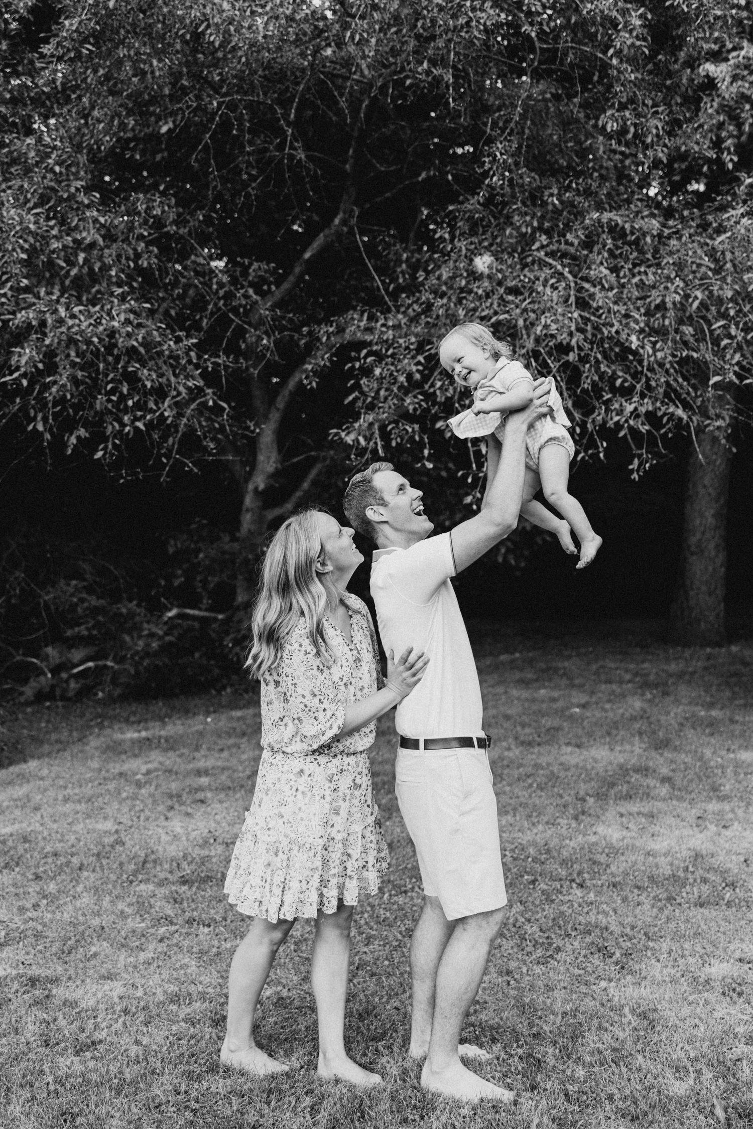 Parents lifting baby in air during playful moment outdoors in black and white photo.