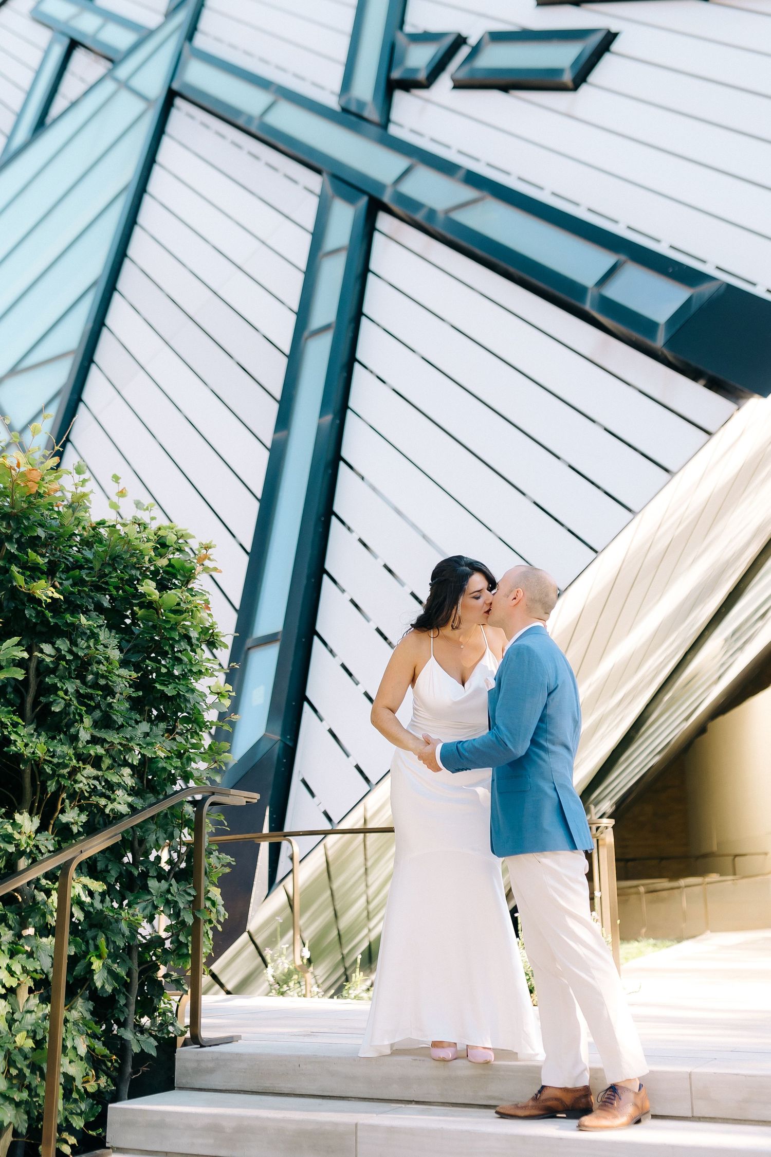 Two people share a romantic moment on the steps of a modern building with a peaked glass roof and white siding.