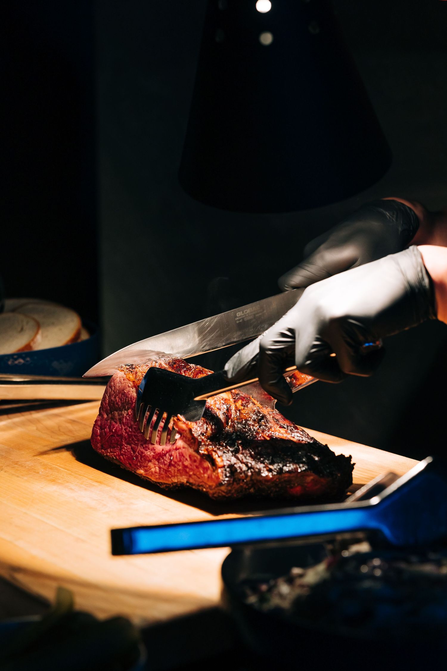 A chef wearing black gloves slices smoked brisket on a wooden cutting board in dramatic lighting.