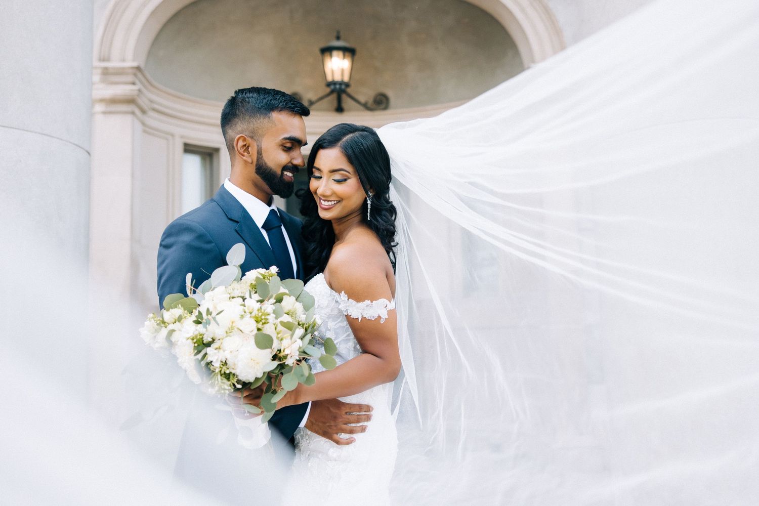 A romantic wedding moment with a flowing veil and white floral bouquet against an elegant arched doorway.