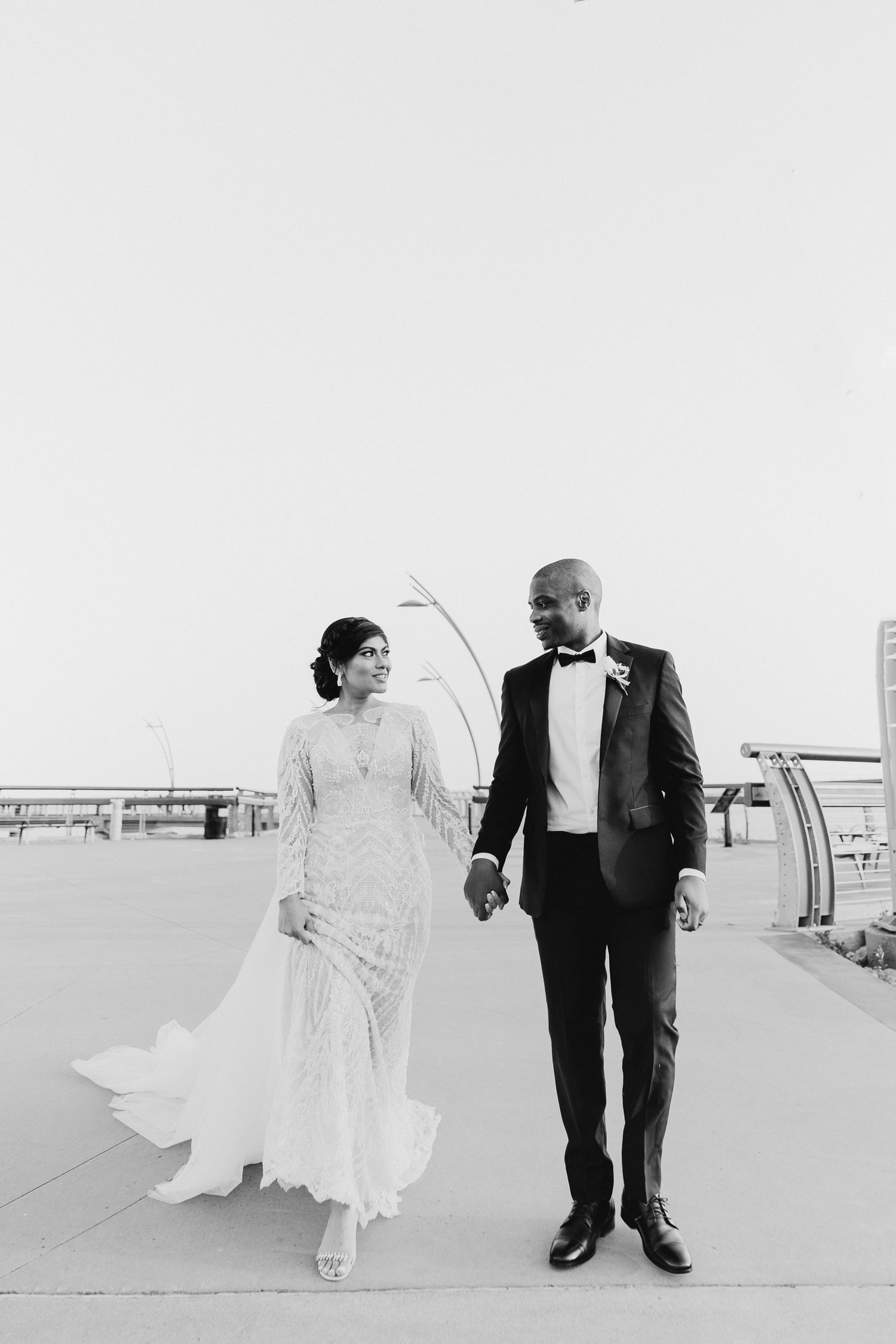 A bride and groom share a romantic black and white moment walking together outdoors in formal wedding attire.