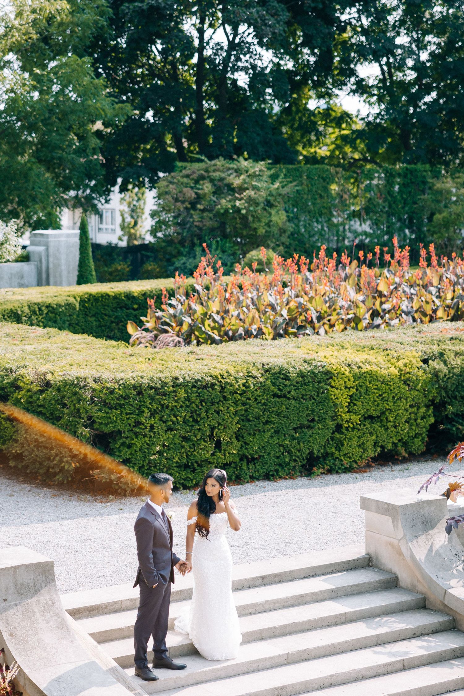 A romantic wedding moment on stone steps surrounded by manicured hedges and colorful garden flowers.