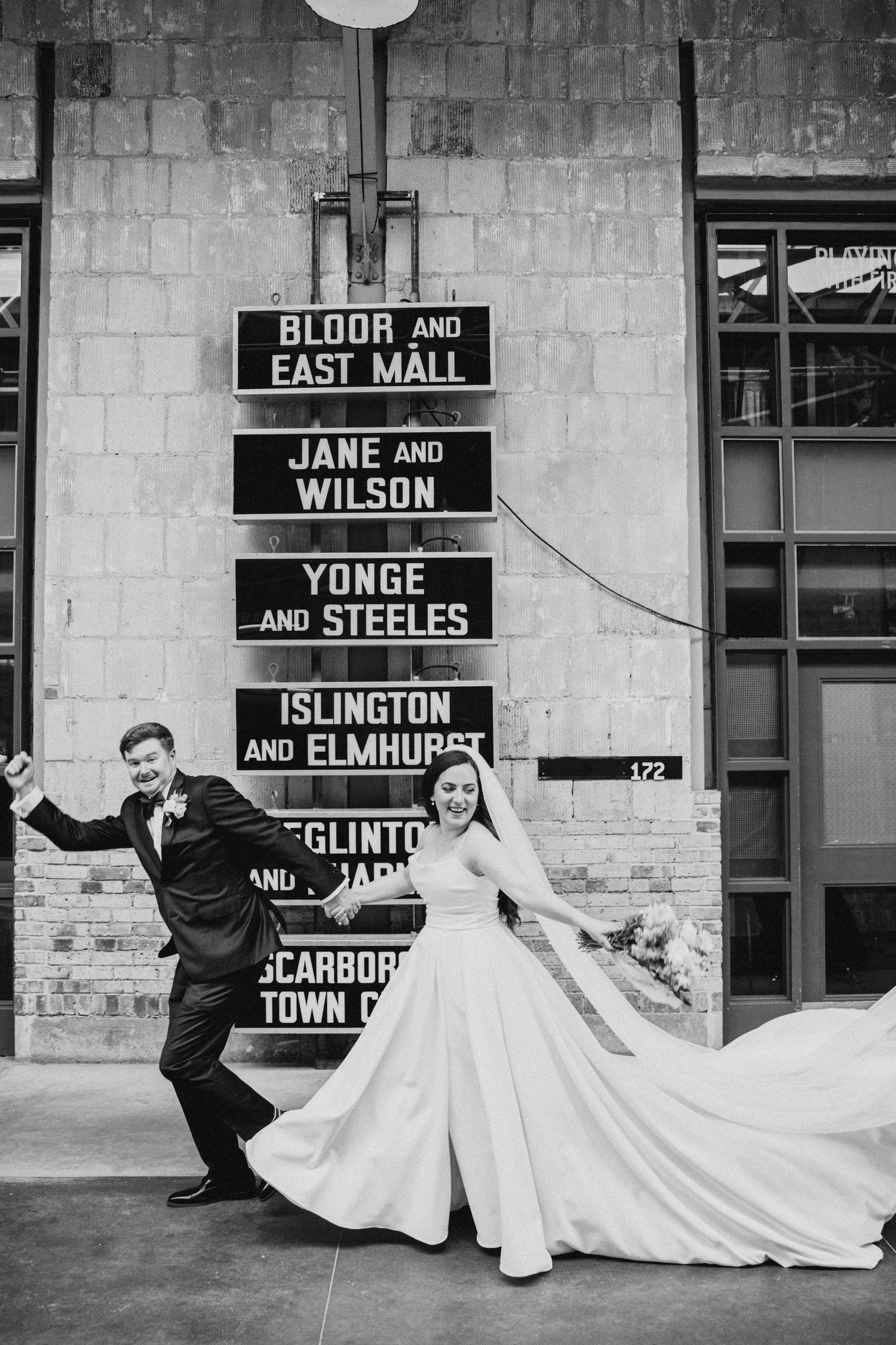 Black and white wedding photo in front of vintage Toronto street signs at a brick wall showing a joyful couple dancing.