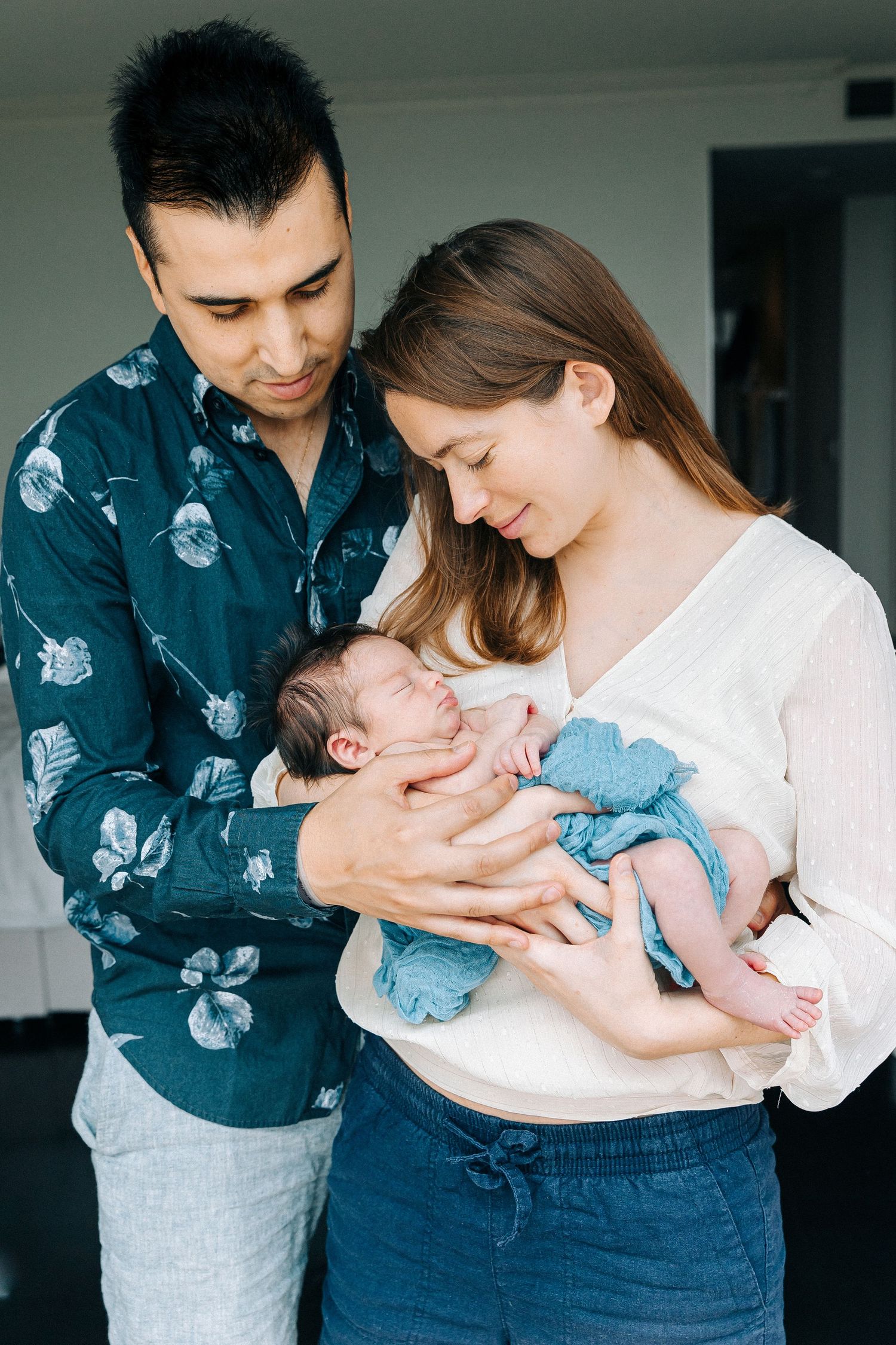 Parents cradling their newborn baby wrapped in a blue blanket in a bright modern room.