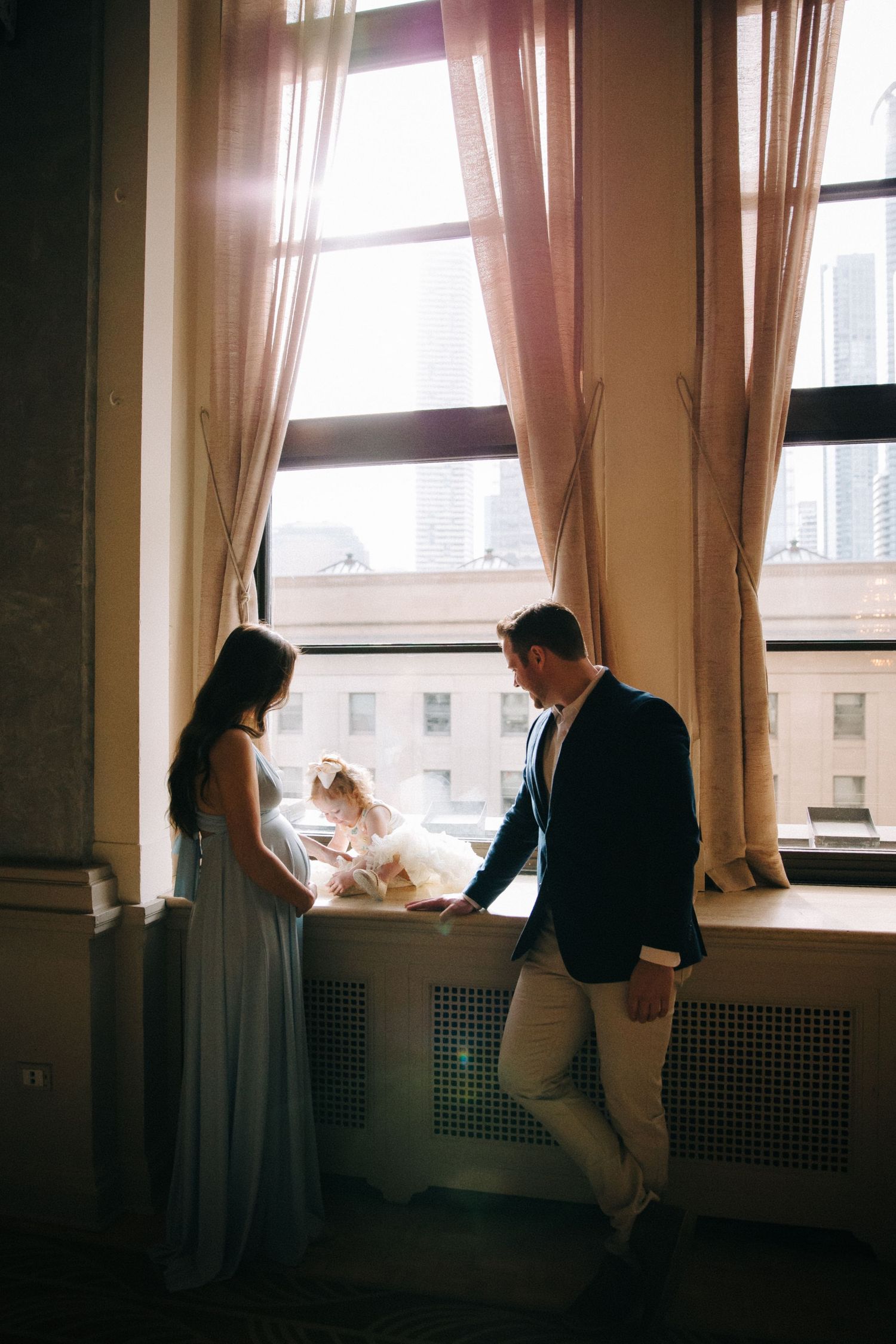 Couple standing by large window with flowing curtains in romantic silhouette.