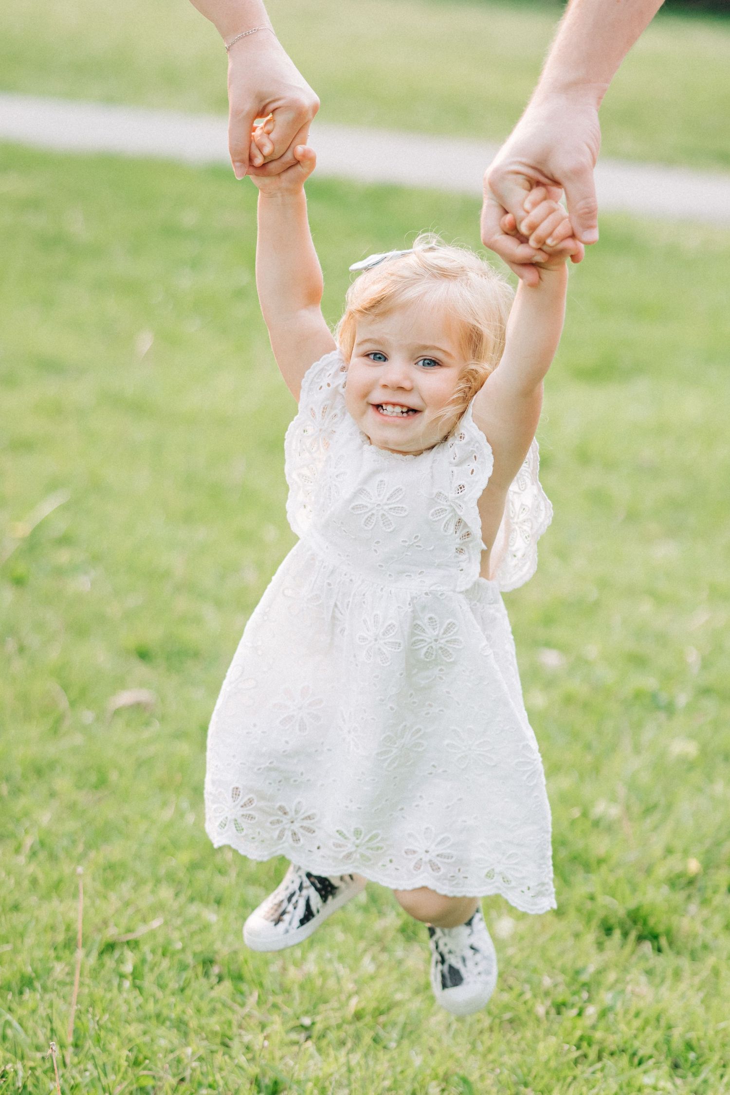 A small child in a white lace dress being lifted up while playing in a grassy field.