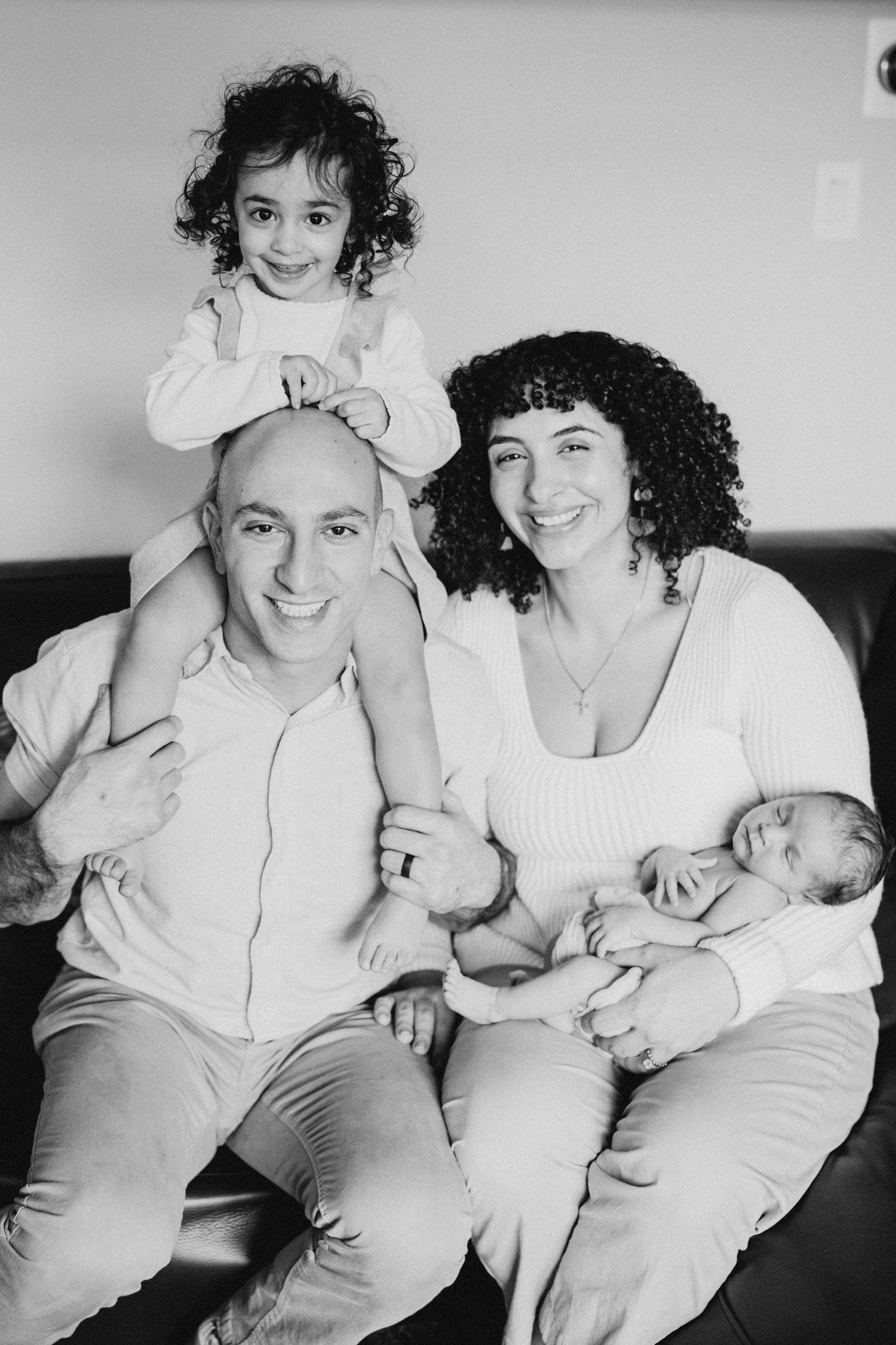 A black and white family portrait showing loving parents with a toddler and baby on a couch.