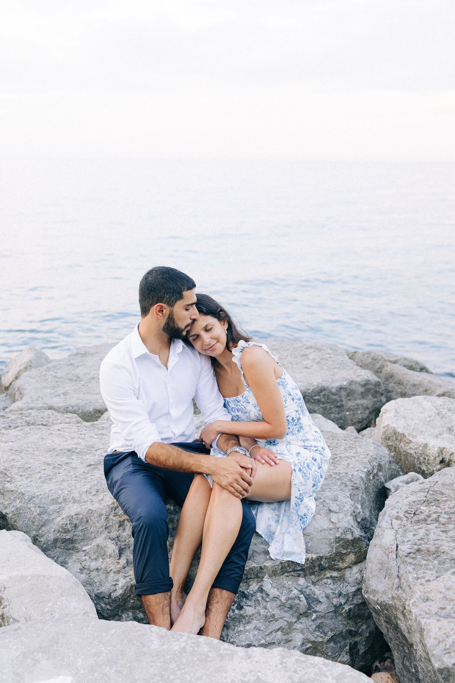 A couple shares an intimate moment while sitting on coastal rocks by the ocean.