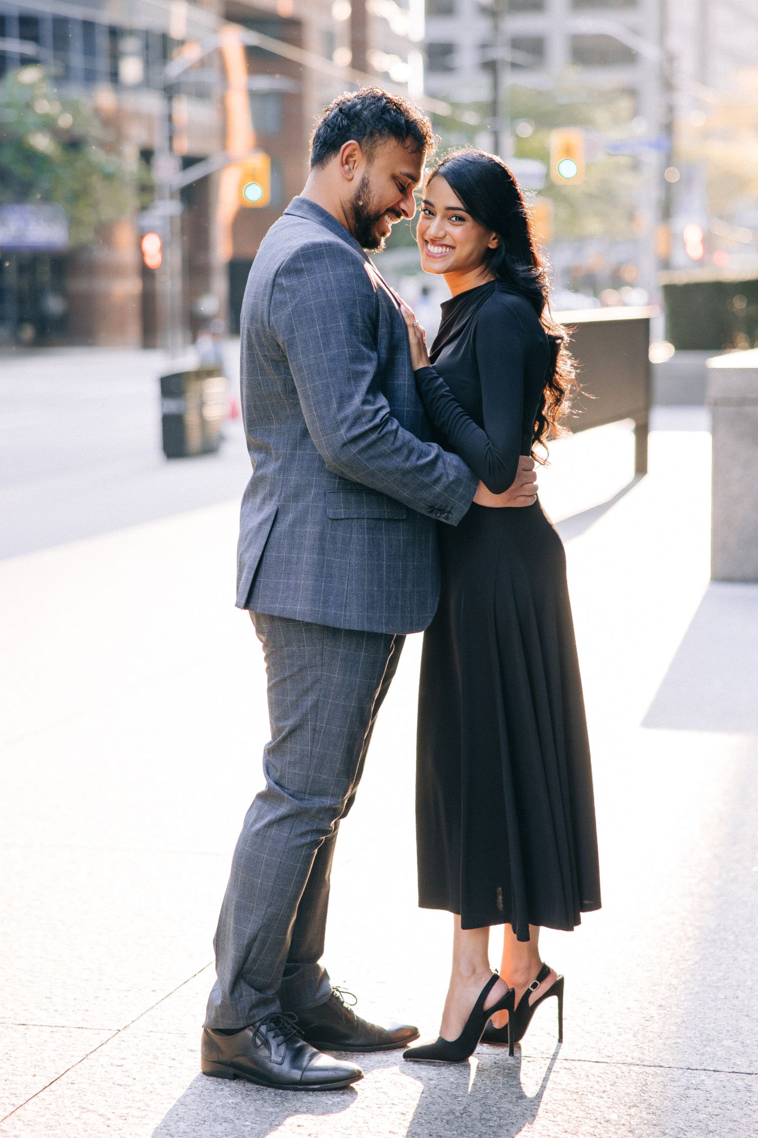 A couple embraces on a city sidewalk, with one wearing a gray suit and the other in a black dress and heels.