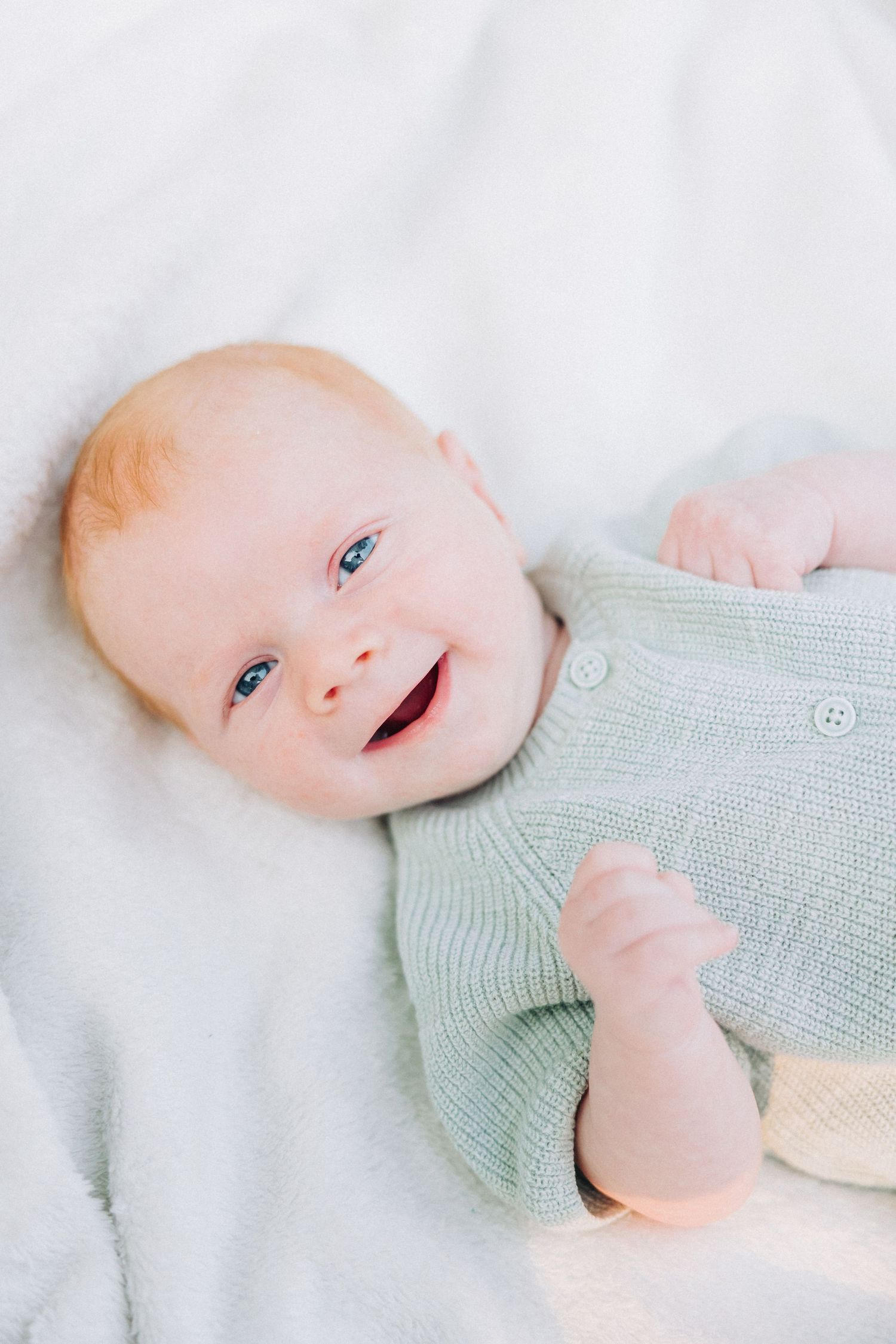A smiling baby in a mint green sweater lying on white bedding.