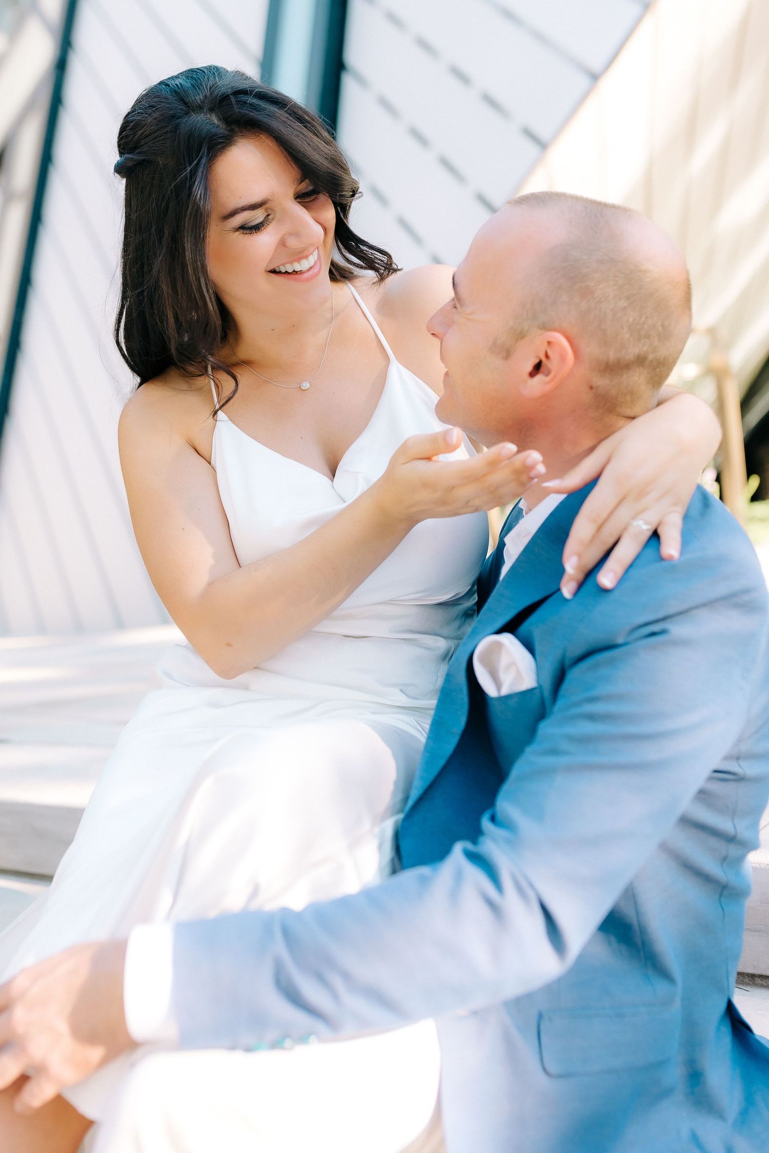 A joyful bride in white dress laughs with groom wearing light blue suit during outdoor wedding photoshoot.