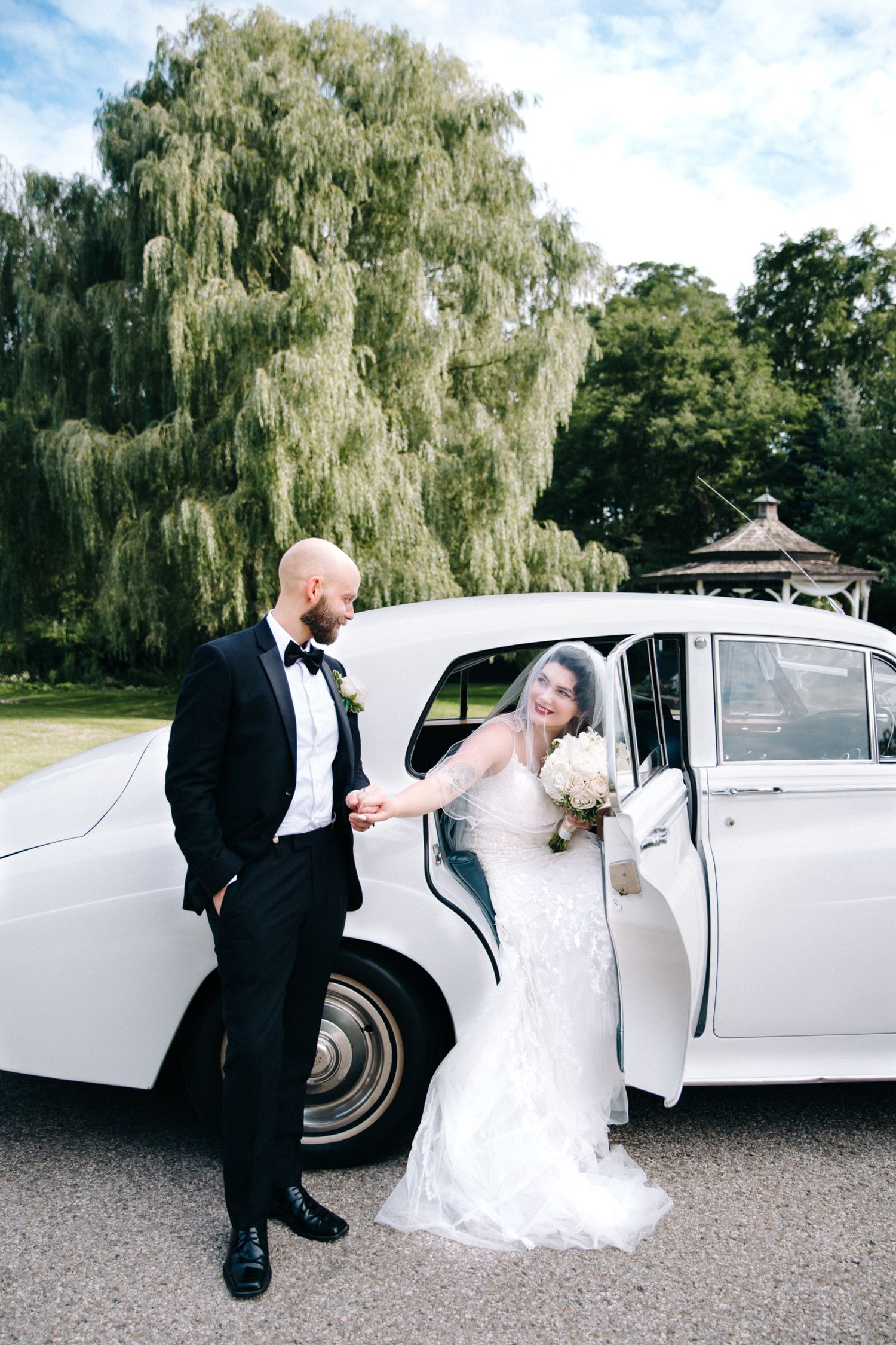 A classic white vintage car parked under a weeping willow tree with a gazebo visible in the background.
