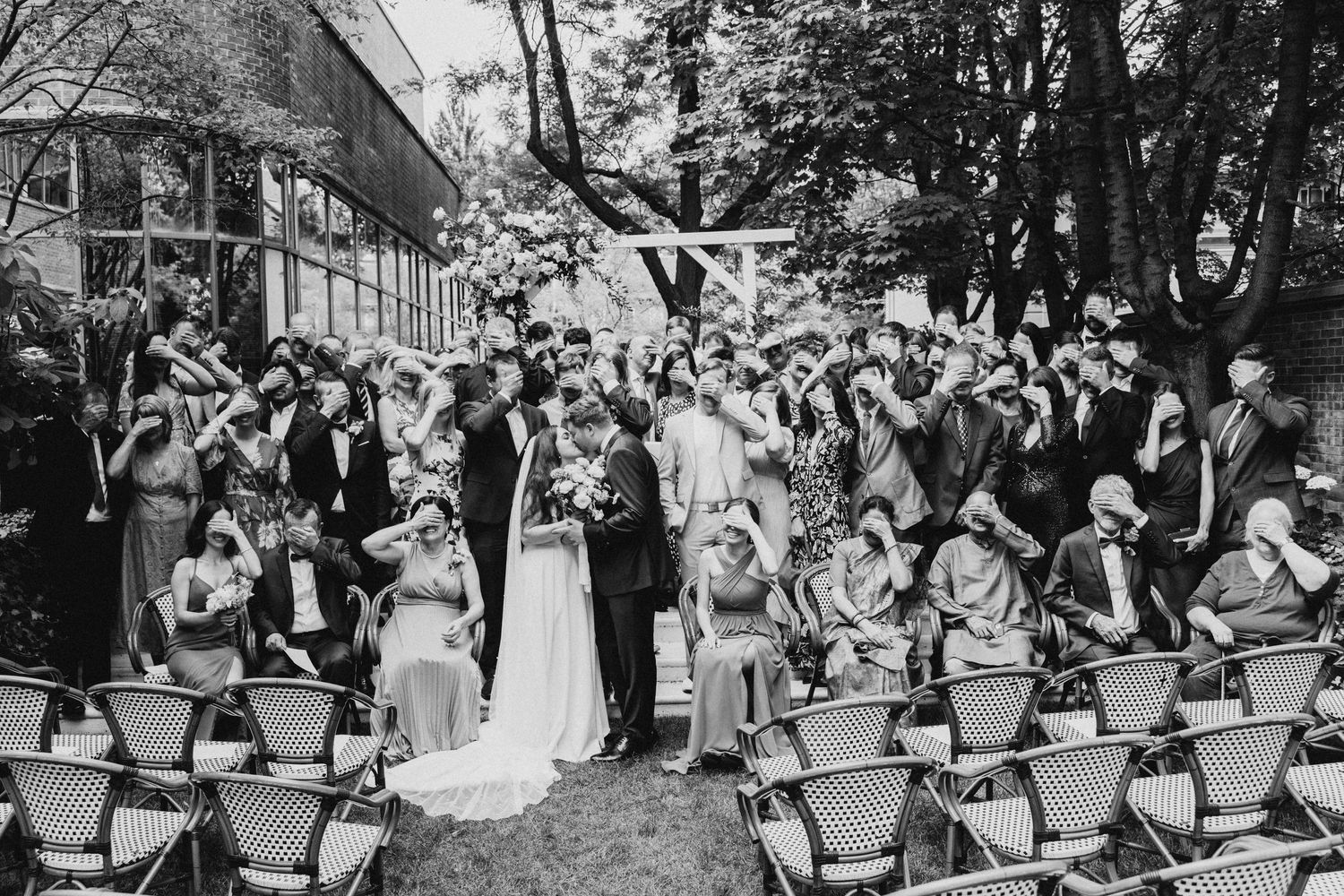 Wedding guests enjoying an outdoor ceremony with rows of folding chairs and tall trees in a vintage black and white scene.