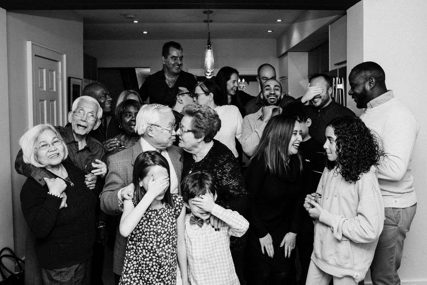 A candid black and white photograph of a group of people laughing together at a casual indoor gathering.
