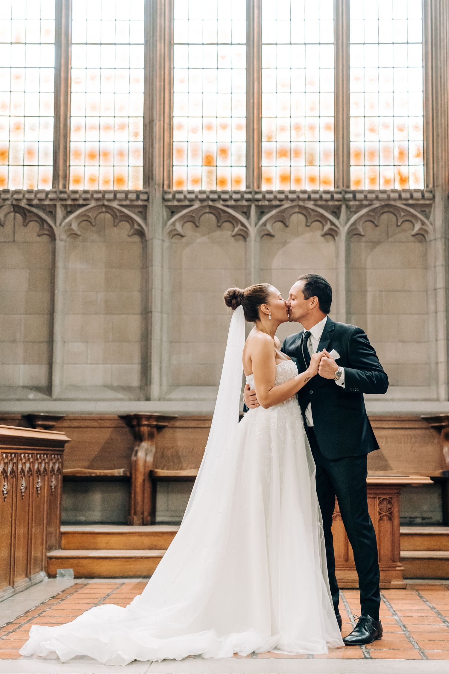 A couple shares a romantic moment in formal wedding attire inside an elegant church with stained glass windows and stone architecture.
