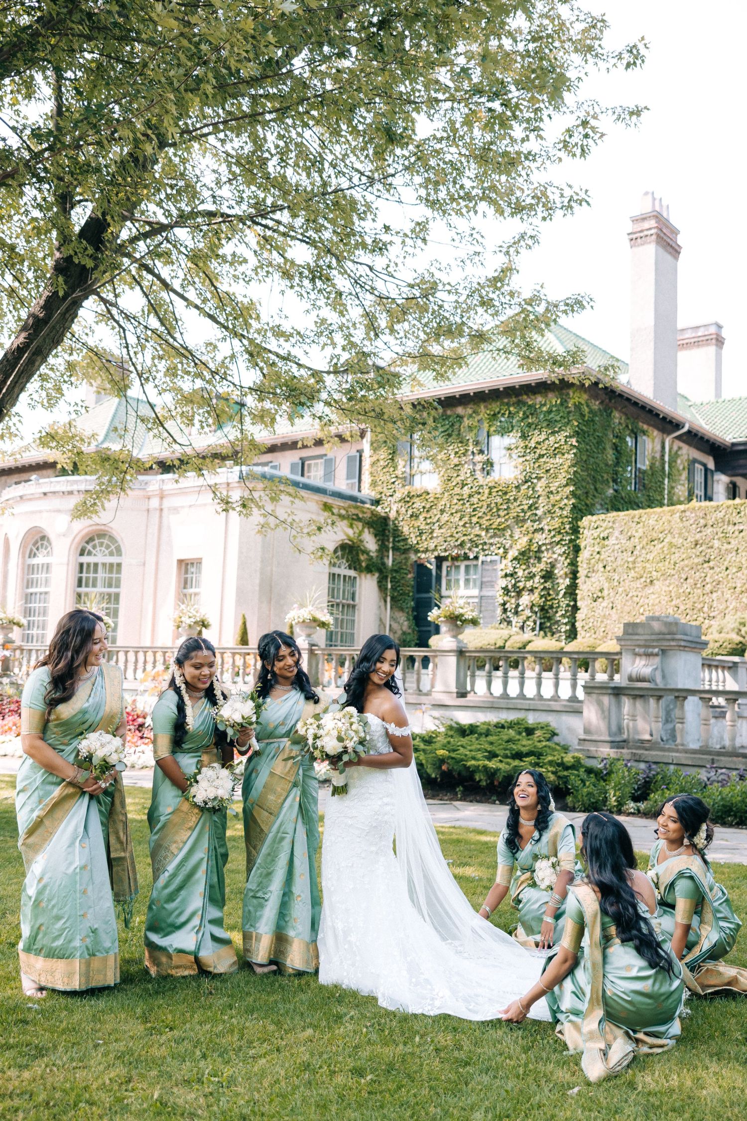 Bridesmaids wearing mint green and gold saris pose on the lawn of an elegant ivy-covered estate mansion.