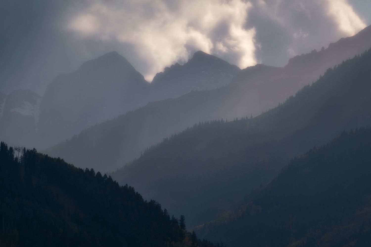 Dramatic mountain landscape with sunlight breaking through clouds over forested peaks in moody atmosphere.