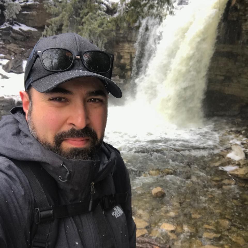An outdoor enthusiast takes a selfie in front of a rushing waterfall with snow visible in the background.